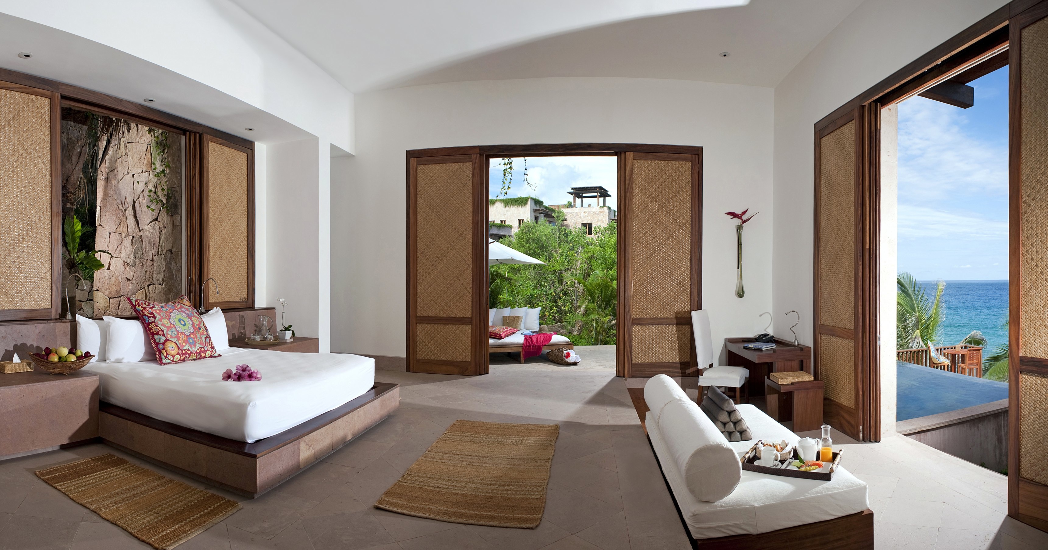 A spacious, tropical-themed bedroom at Imanta Resort features a bed, couch, and open doors revealing a patio with greenery and an ocean view. The room boasts natural wood accents and clean, minimalist decor, creating a serene spa-like atmosphere.