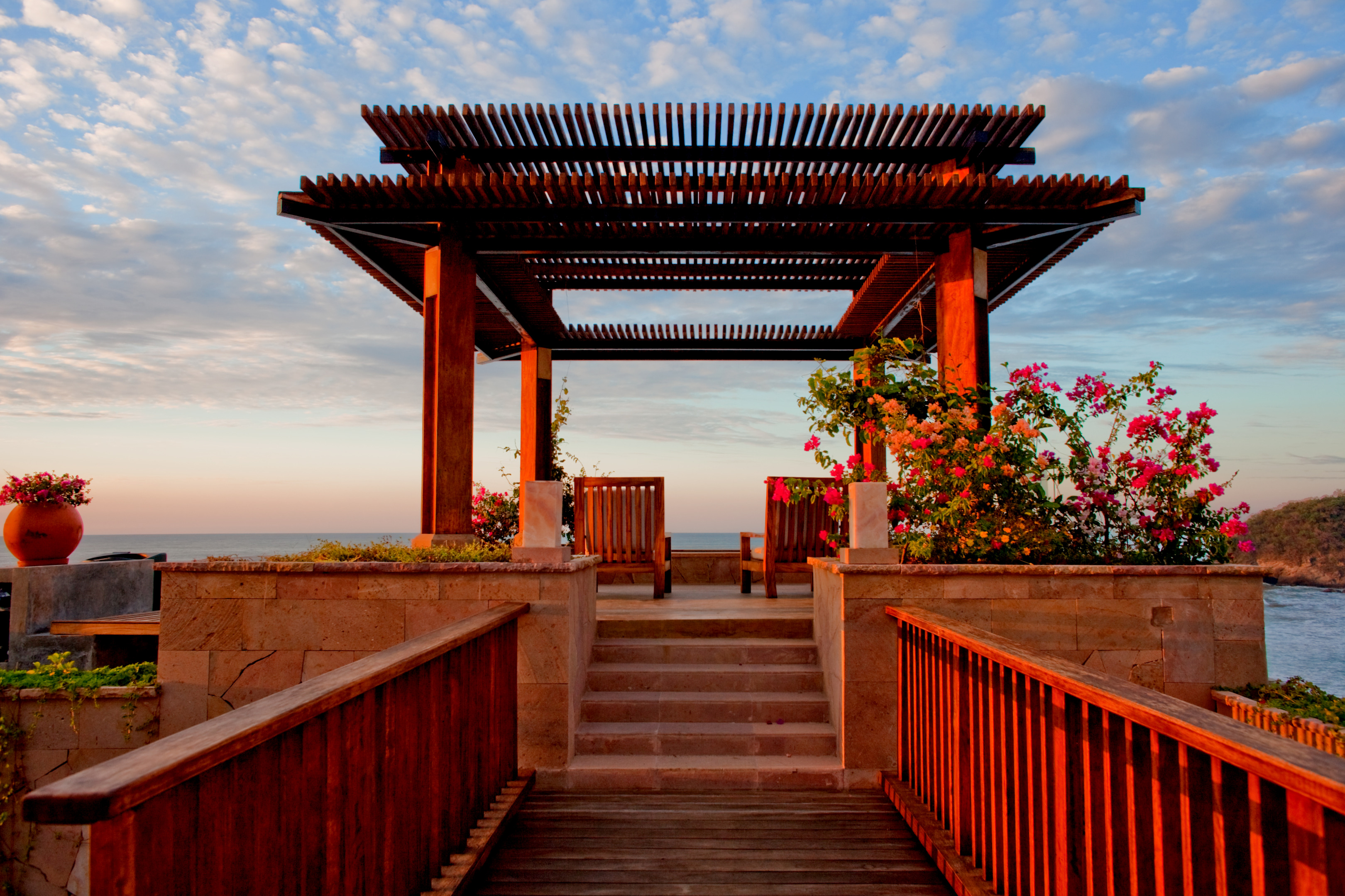 A wooden gazebo with flowers offers ocean views under a partly cloudy sky at Imanta Resort, accessible via a wooden path and steps, perfect for relaxation before or after a spa session.