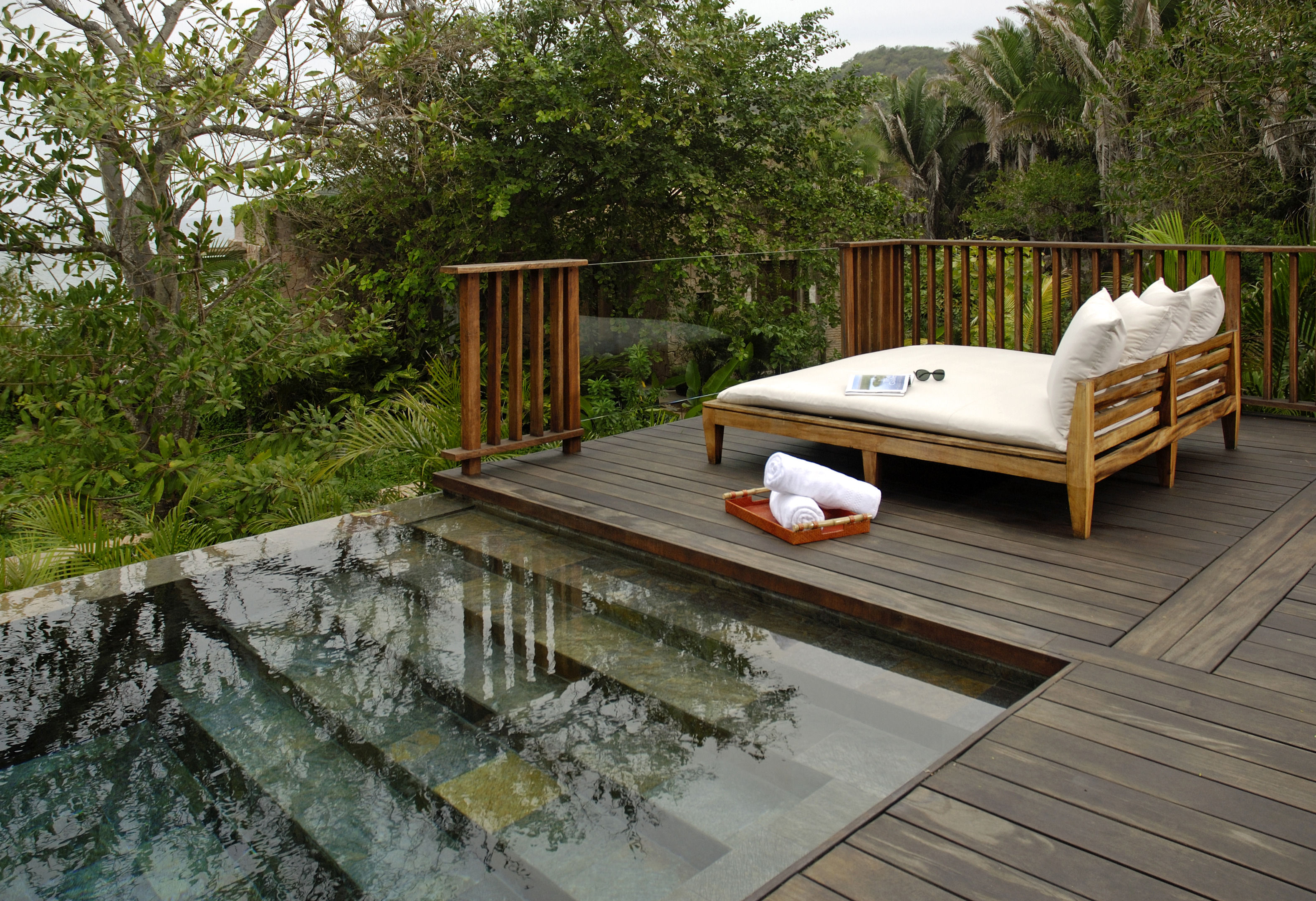 Outdoor deck with a wooden daybed featuring white cushions and a rolled towel beside an infinity pool at Imanta Resort. Surrounded by lush green tropical vegetation, it offers a perfect spot to unwind after a spa treatment.