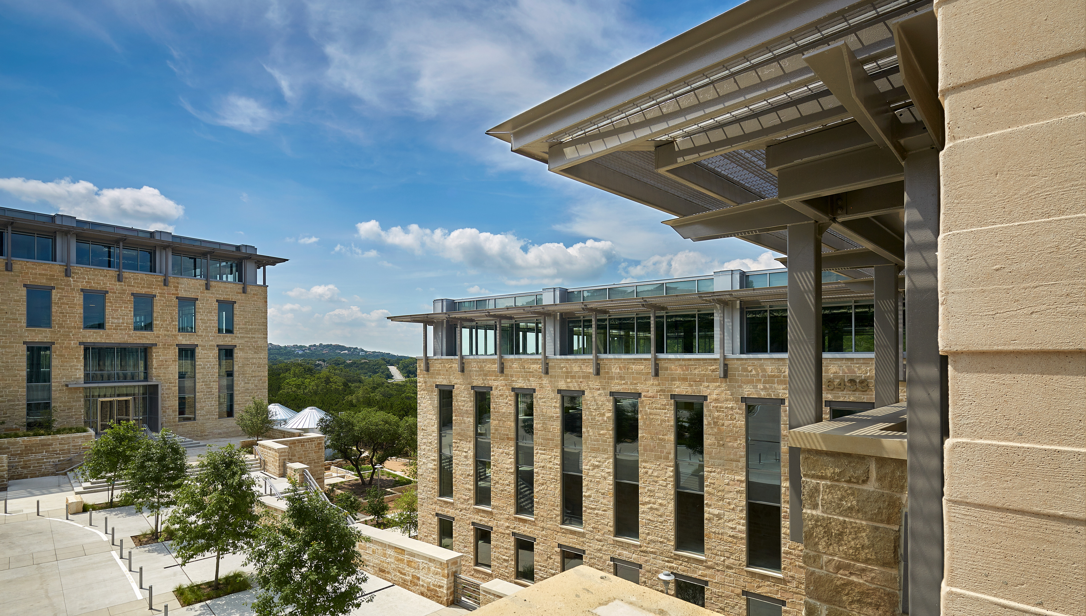 Modern office buildings with stone facades, large windows, and exposed metal framework stand against a backdrop of trees and hills under a clear blue sky, resembling a meticulously planned auto draft.