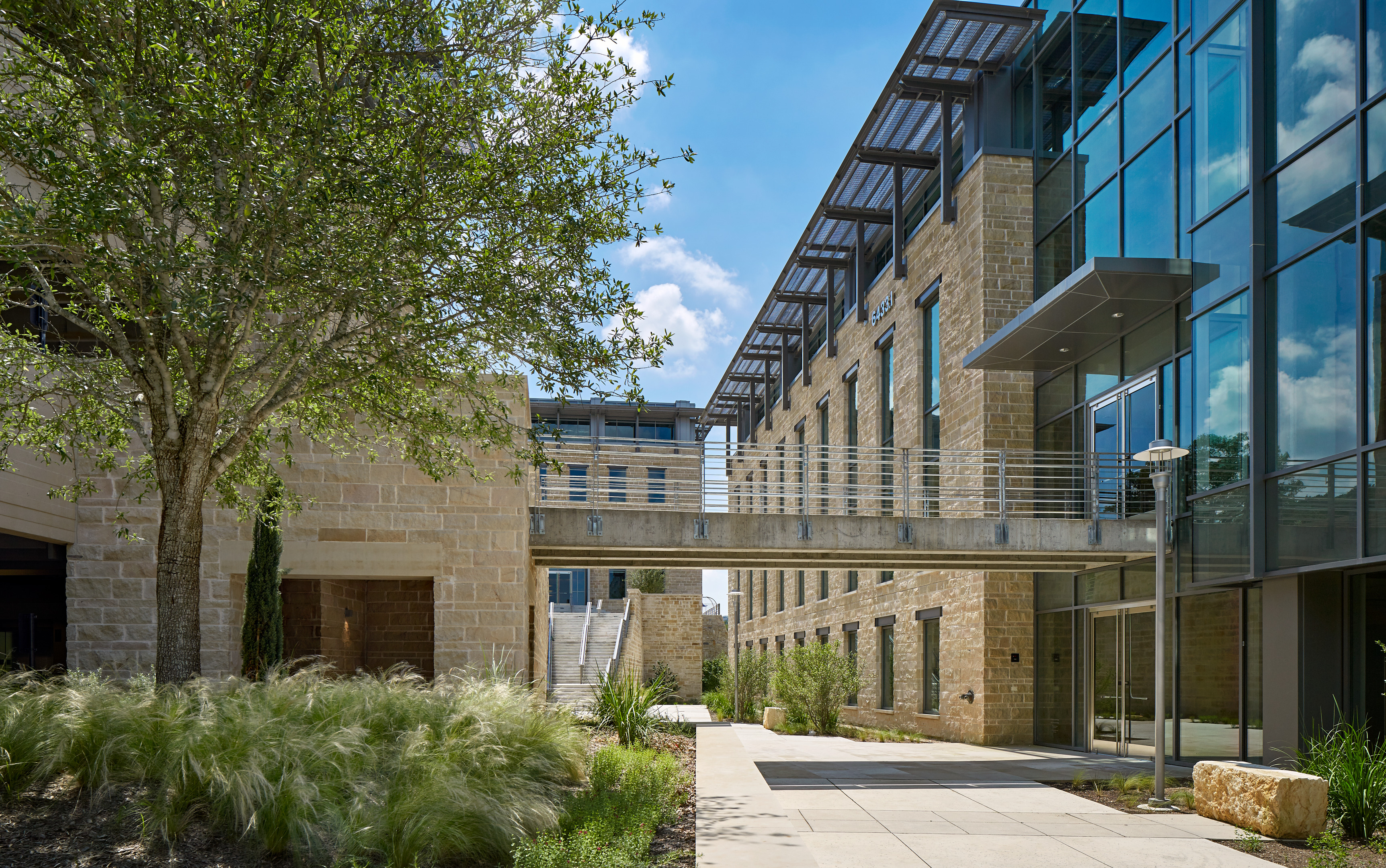 A modern building with stone walls and large glass windows, featuring a skybridge connecting to another section over a neatly landscaped walkway, seems almost like an auto draft from an architect's imagination.