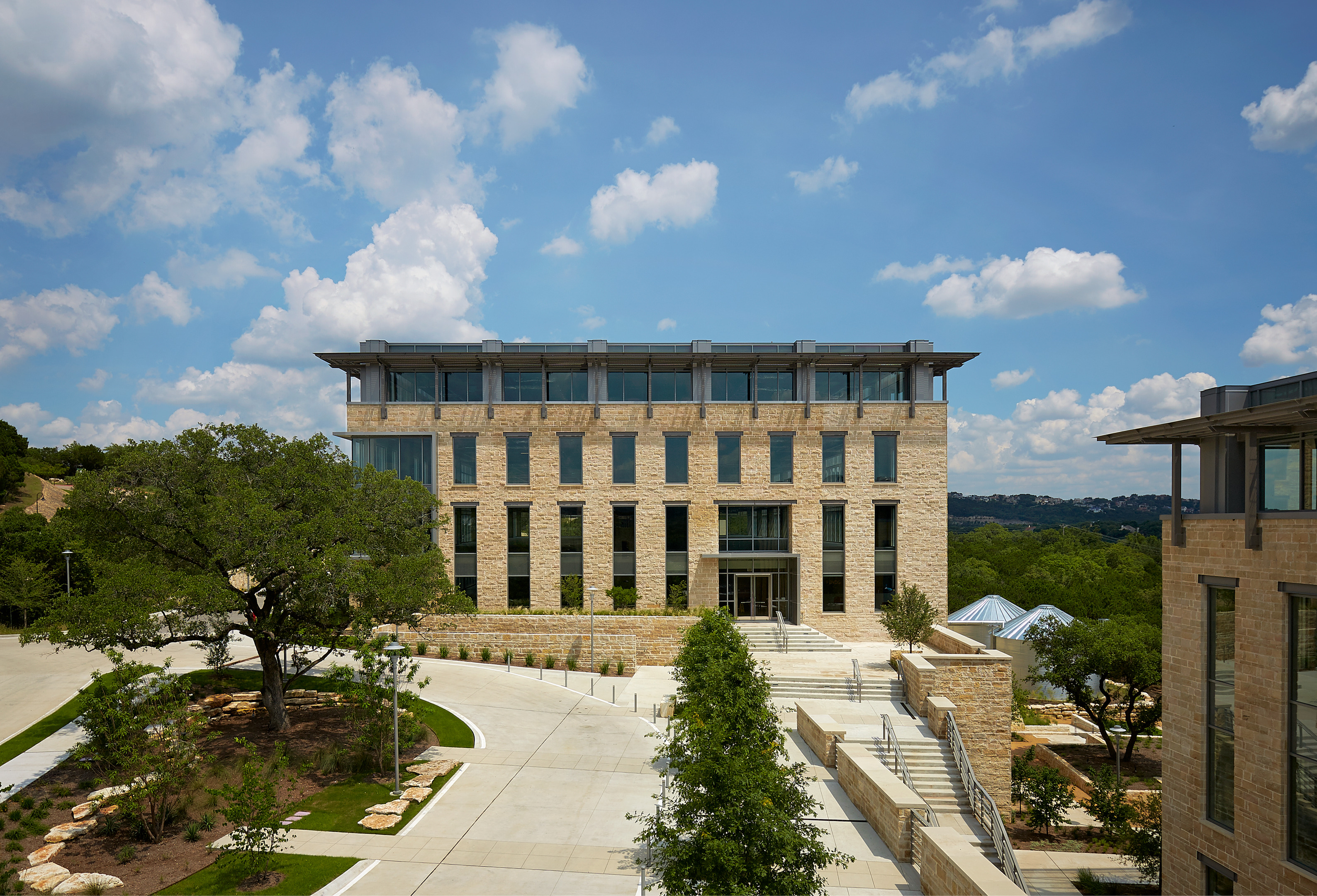 A modern, multi-story building with large windows and stone facade stands impressively, set against a backdrop of trees and a blue sky with scattered clouds. The foreground features landscaped greenery, a concrete pathway, and incorporates an auto draft system for optimal efficiency.