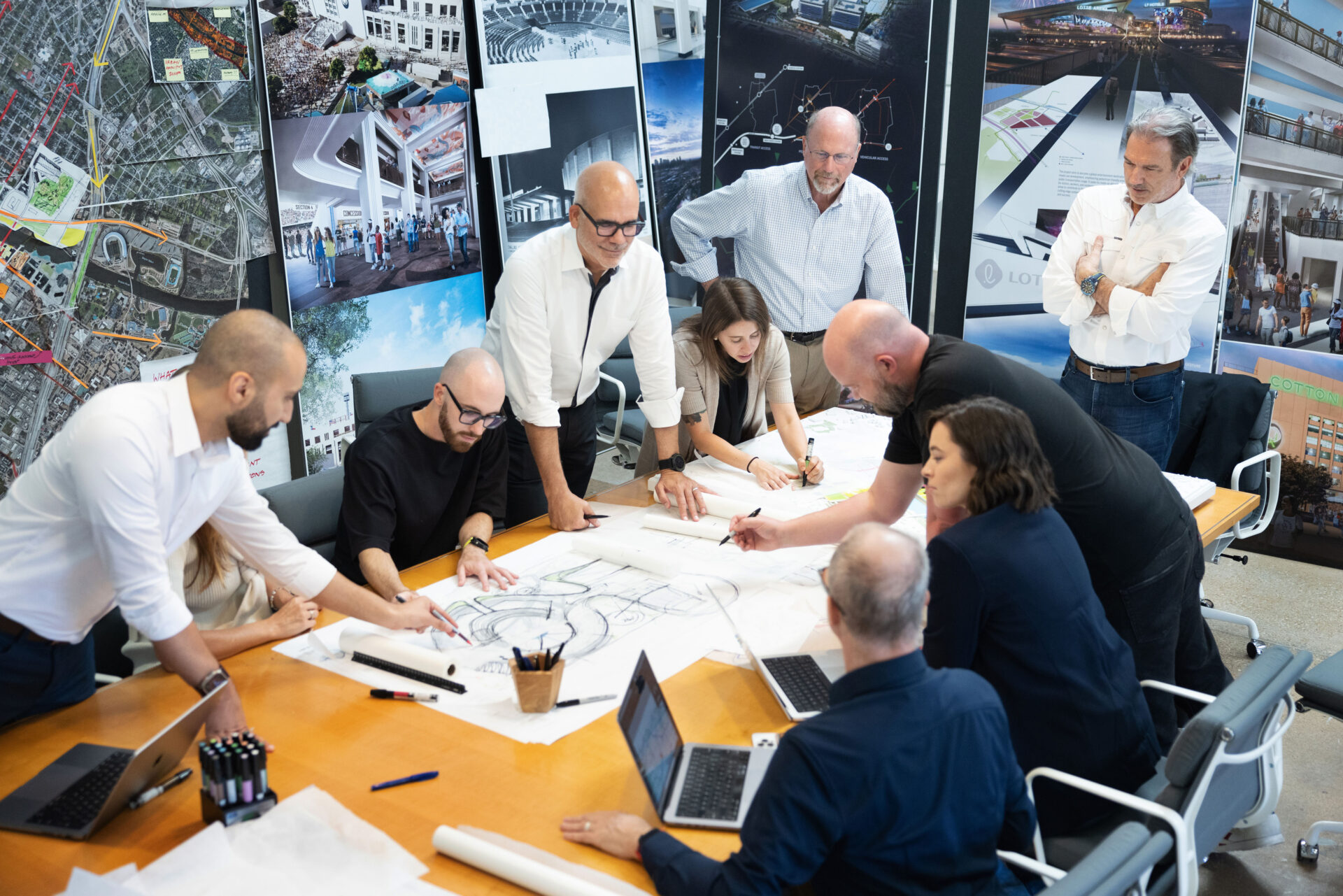 A group of people are gathered around a table covered with blueprints and papers, discussing and pointing at the documents. Several laptops are open on the table, surrounded by design posters on the walls, reflecting a collaborative culture.