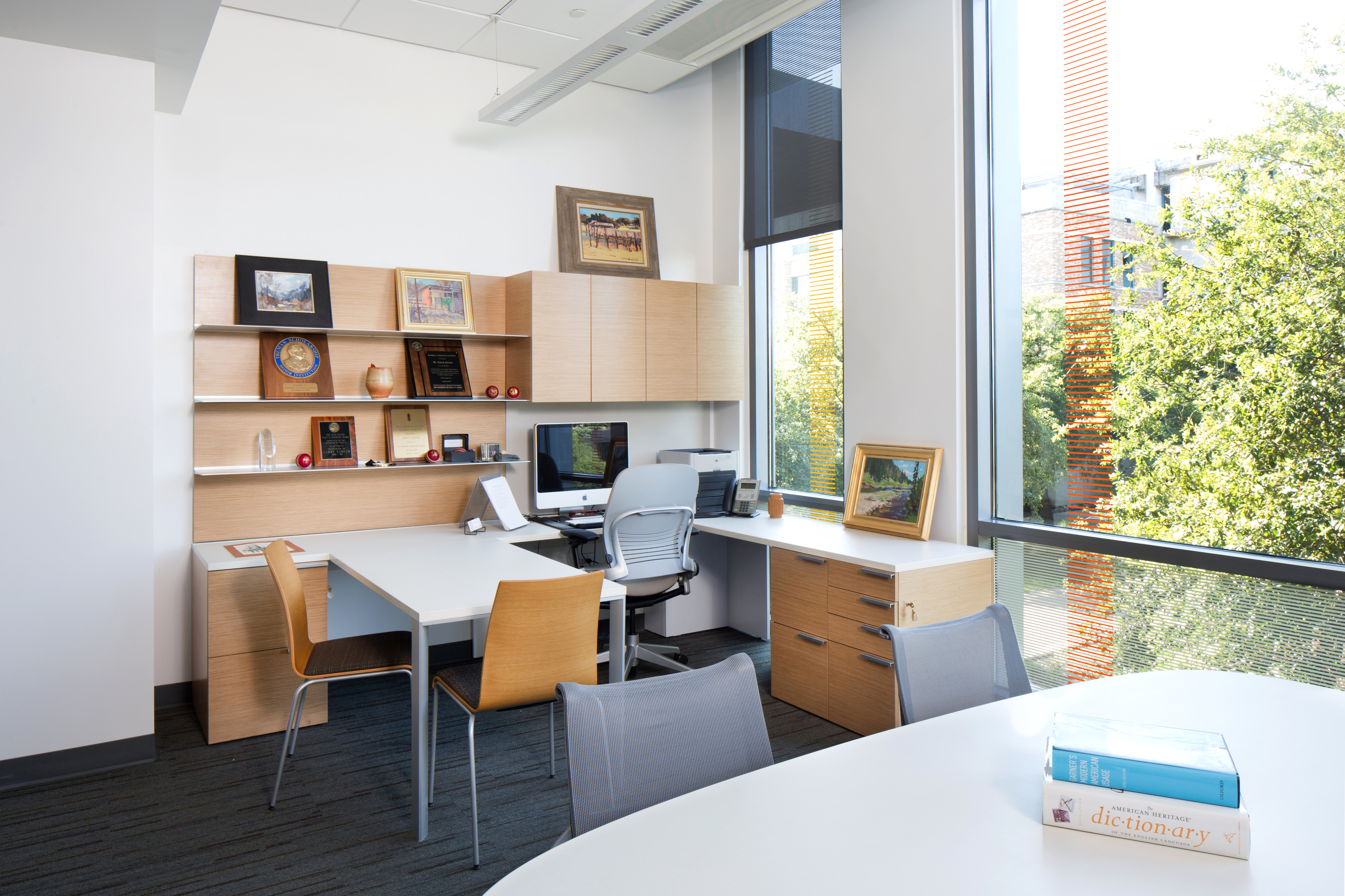 A modern office in Patton Hall with a large window, a desk with a computer, a chair, wooden cabinets, a smaller table with another chair, and in the foreground, a table adorned with books including a dictionary.