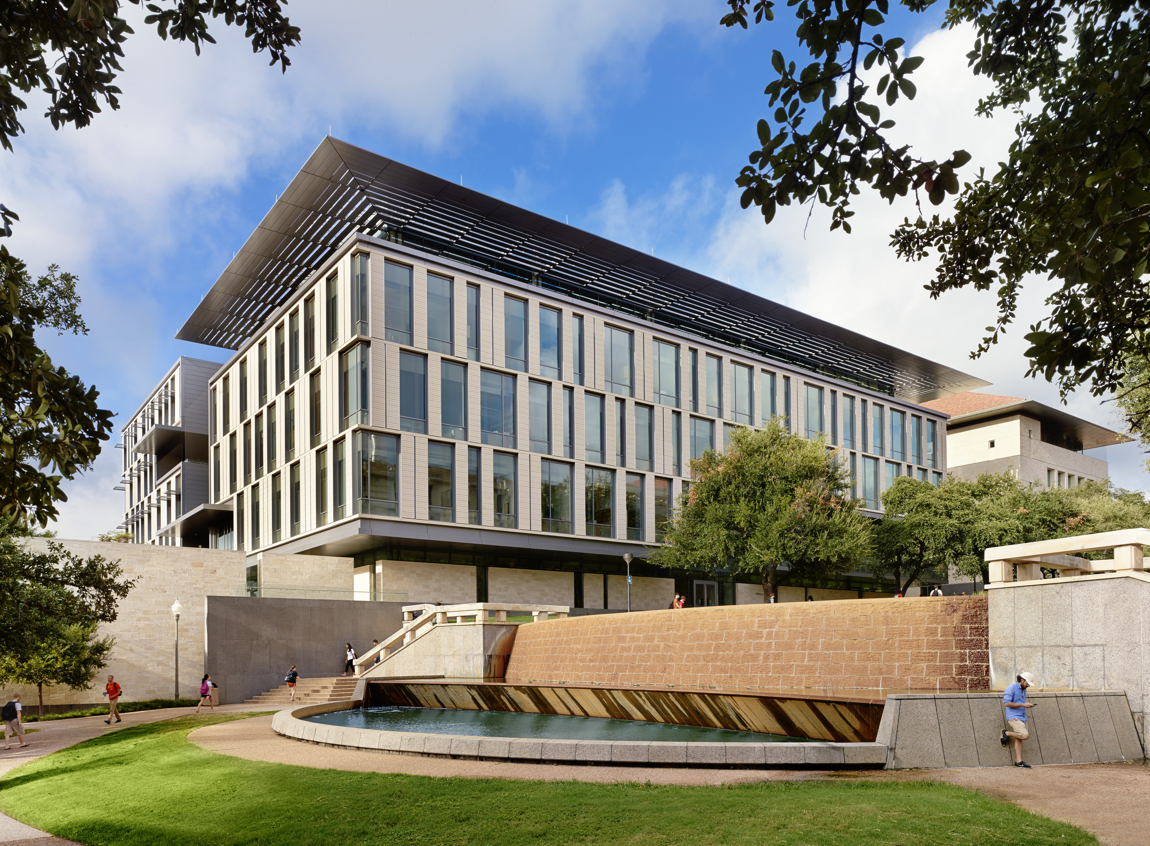 A modern multi-story building with large windows, surrounded by trees, an inclined grassy area, and a cascading water feature in the foreground. People are walking near Patton Hall at the University of Texas.