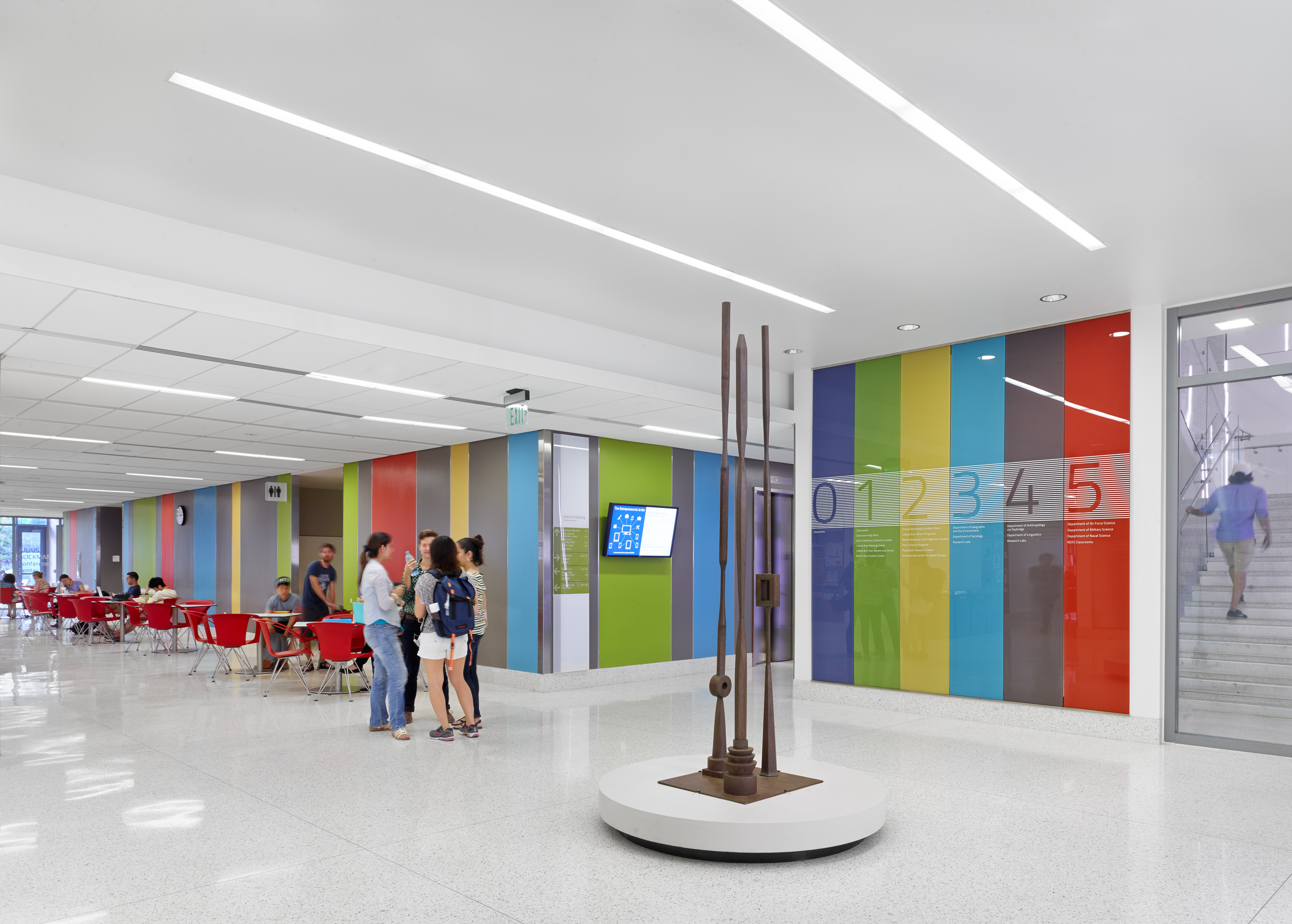 A modern indoor space at Patton Hall with colorful wall panels, red chairs, and tables to the left. A metal sculpture stands in the middle while people walk and converse. Stairs leading to more of the University of Texas campus are visible in the background to the right.