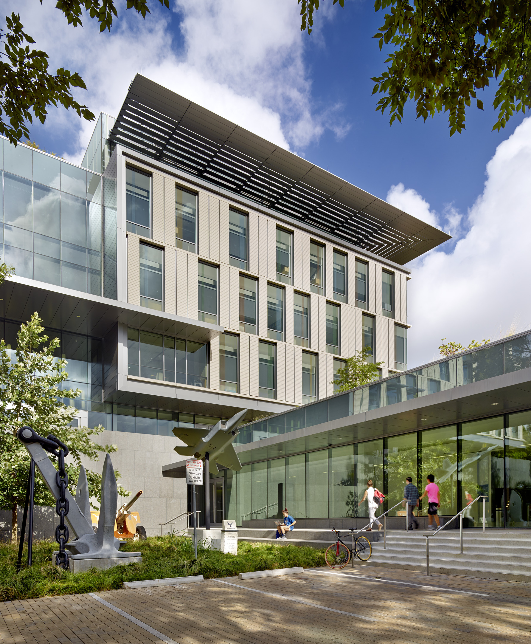 The modern Patton Hall at the University of Texas features a sleek glass and metal facade surrounded by trees. People are walking and sitting near the entrance, where a large anchor sculpture stands prominently in front of the building.