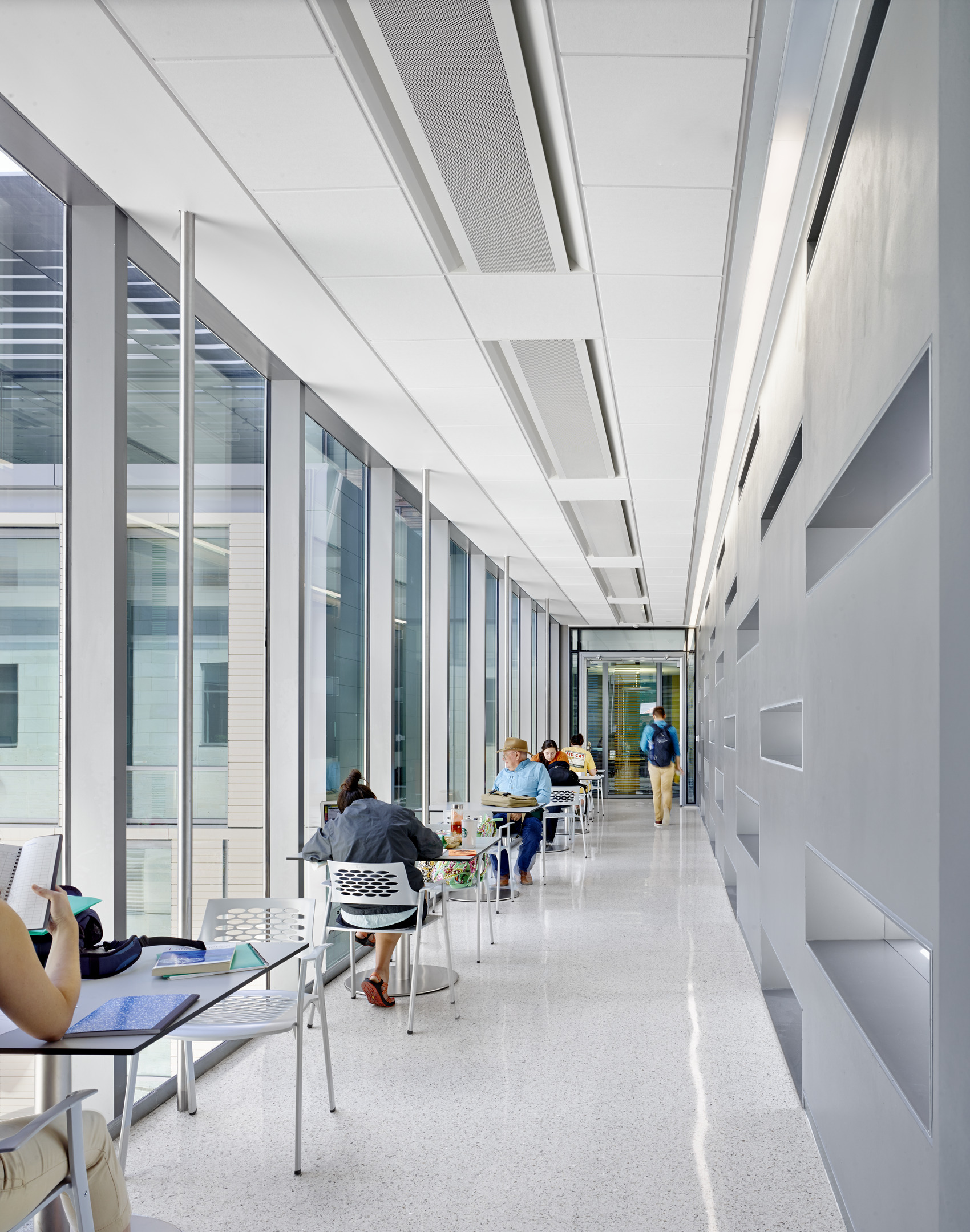 People are working at tables in a long, modern hallway with large windows and a light color scheme, reminiscent of the inviting atmosphere found in Patton Hall at the University of Texas.