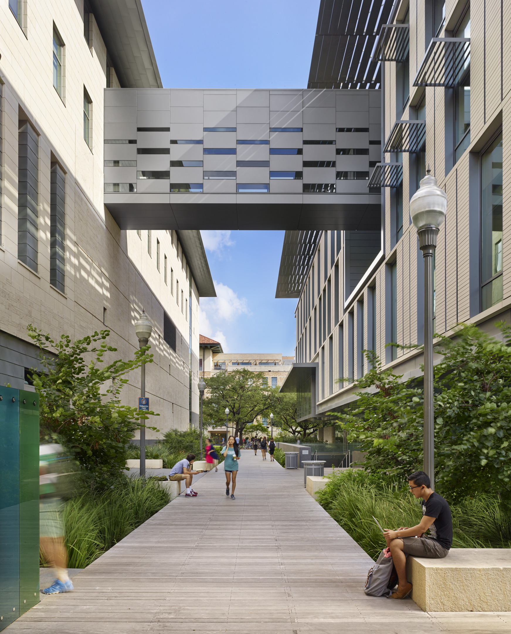 A modern, elevated walkway connects two buildings on the University of Texas campus. People walk and sit in the landscaped pathway below Patton Hall on a sunny day.