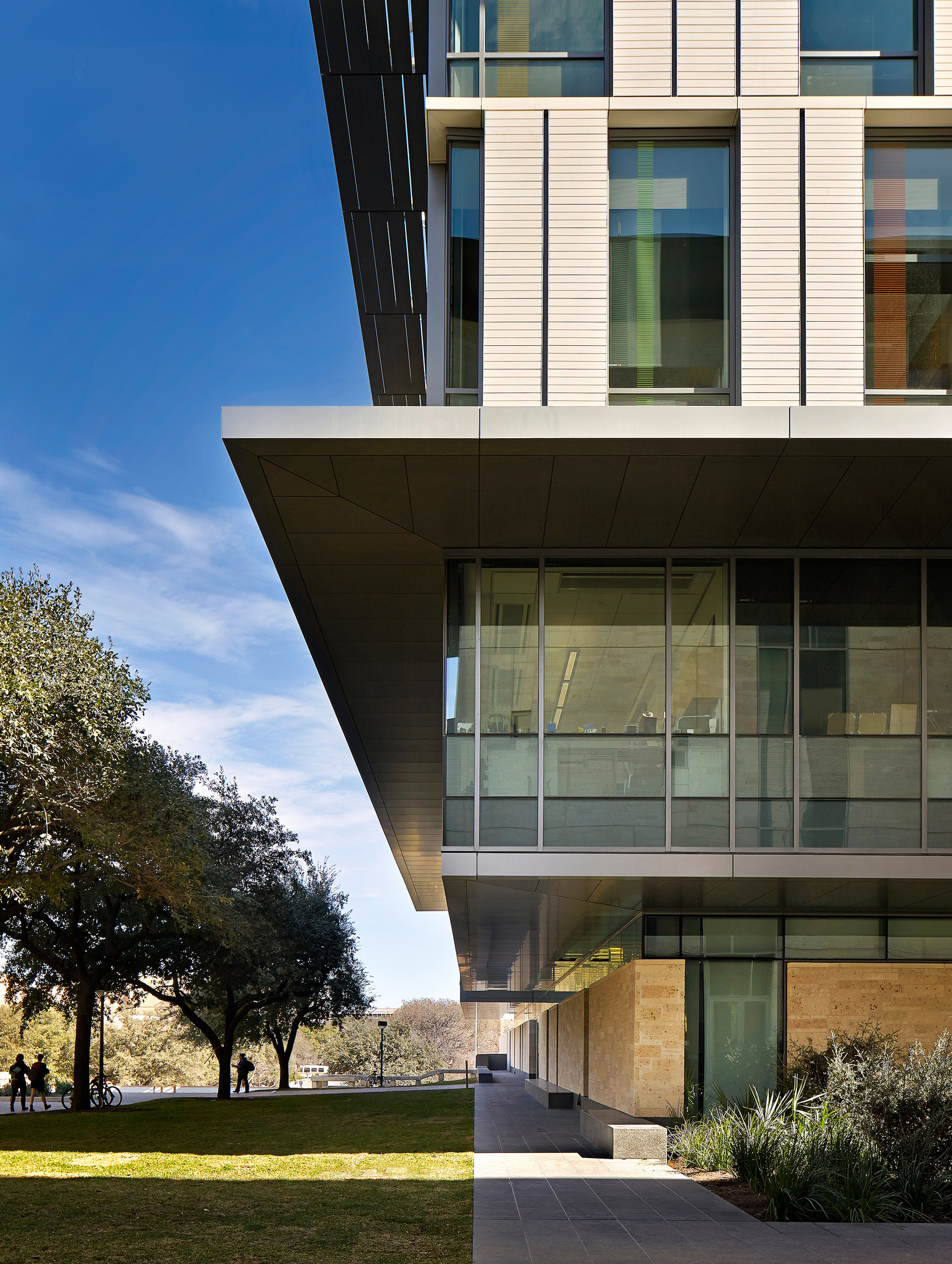 Patton Hall at the University of Texas is a modern building with large windows and an overhanging structure under a clear blue sky, surrounded by trees and green grass. People and bikes are visible in the distance.