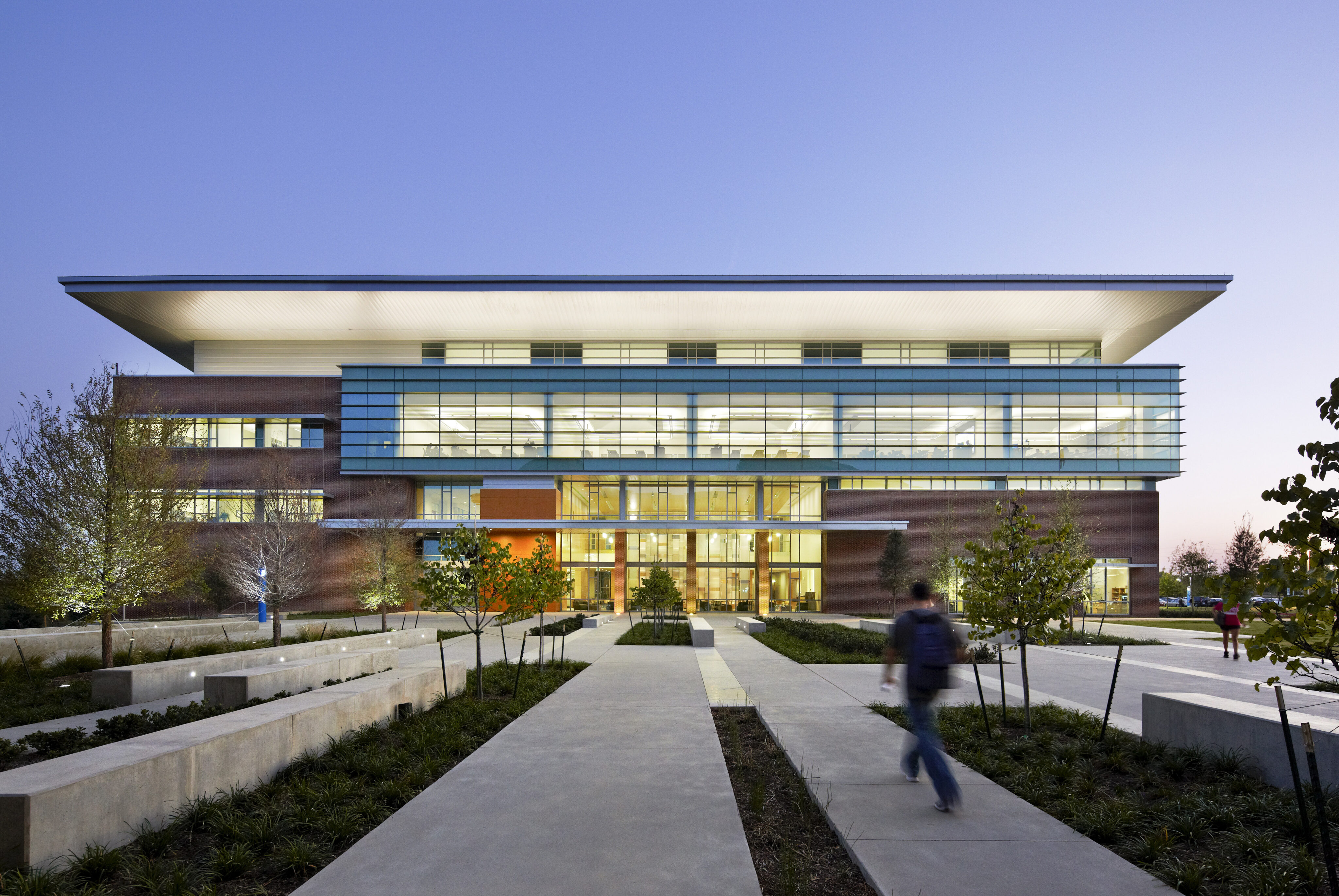 A modern, multi-story glass and brick building with a roof overhang, surrounded by landscaped walkways and greenery; a person walks towards the entrance at dusk. This could be Founders Hall at the University of North Texas, UNT Dallas.