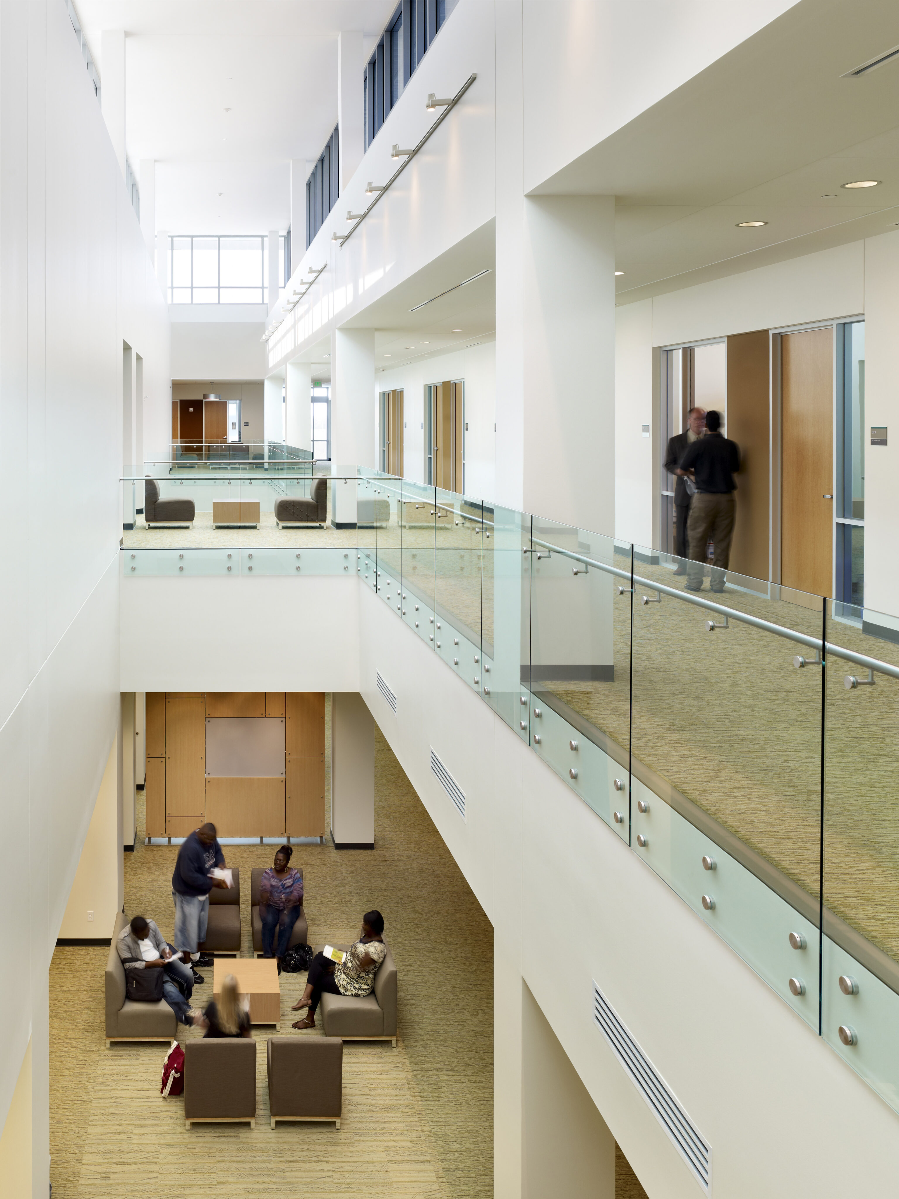 A modern indoor atrium with glass railings at Founders Hall, University of North Texas, shows people in conversation on the ground floor seating area. Two individuals can be seen walking on the upper level.