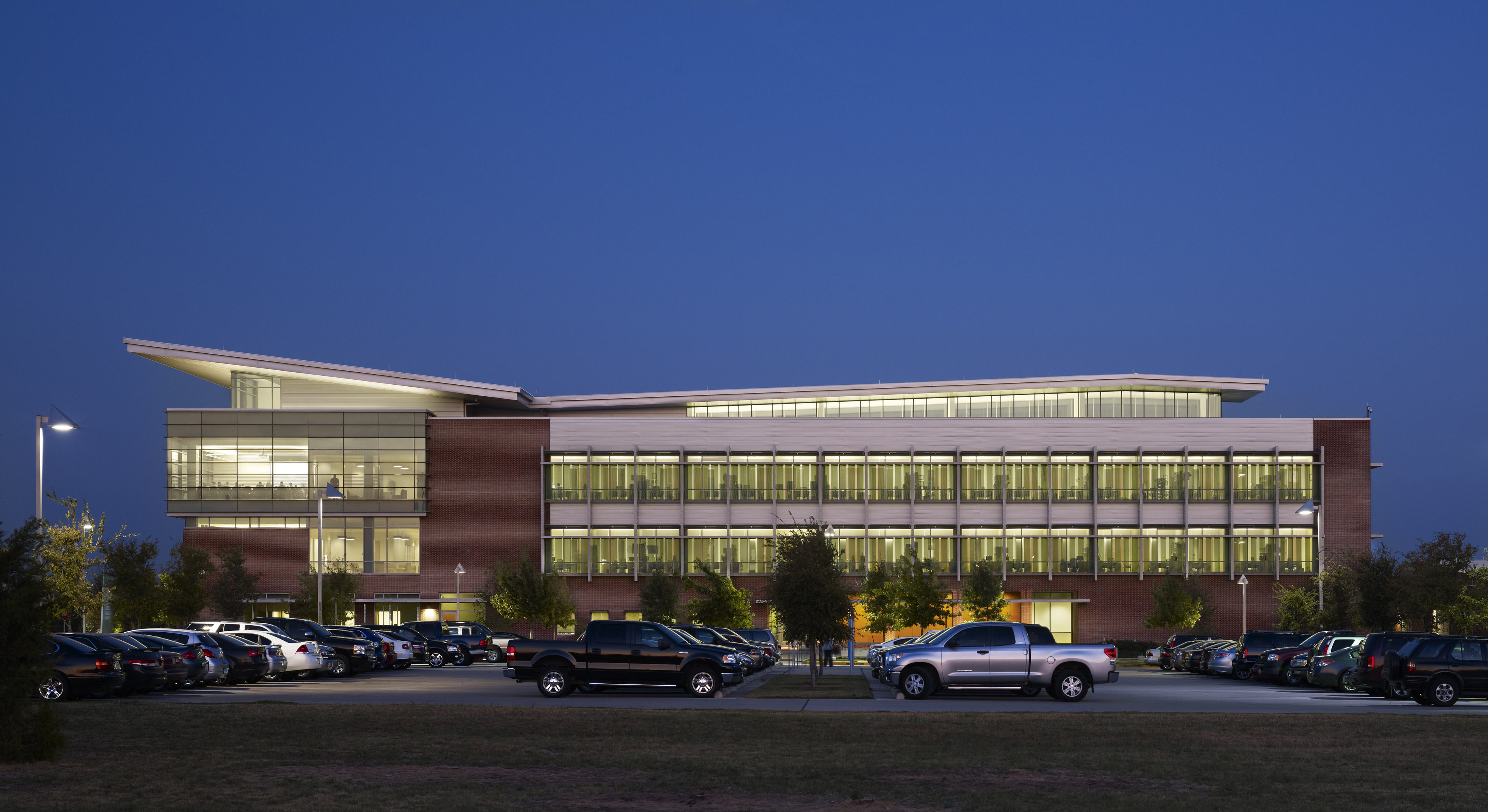 A modern office building with large windows, known as Founders Hall at the University of North Texas in Dallas, is seen at dusk. The parking lot in front is filled with cars, and the building is illuminated from within, contrasting with the darkening sky.