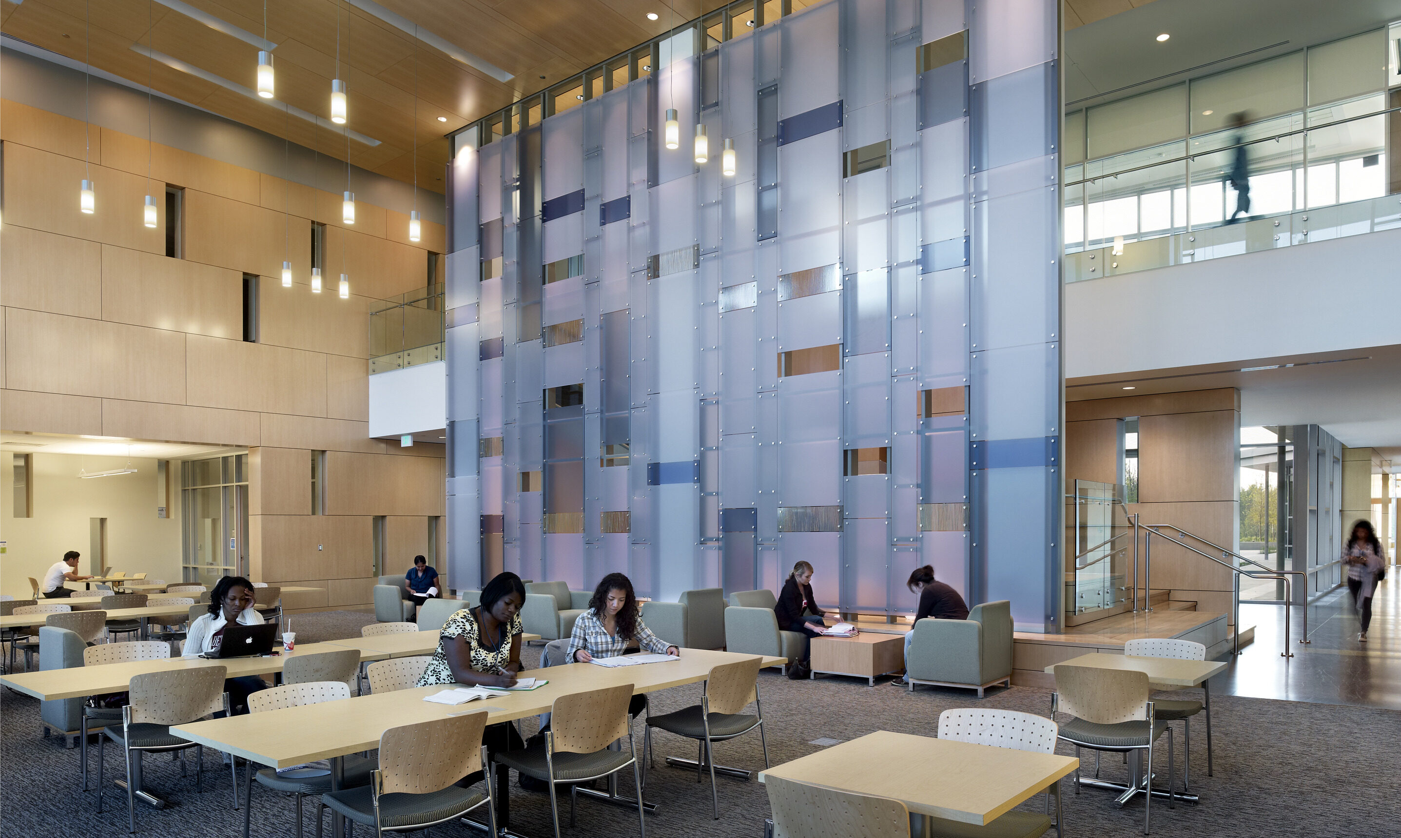 Spacious modern lounge area with high ceilings, hanging lights, wooden accents, and people seated at tables and chairs, engaging in various activities like reading and using laptops; a typical afternoon scene in Founders Hall at the University of North Texas in Dallas.