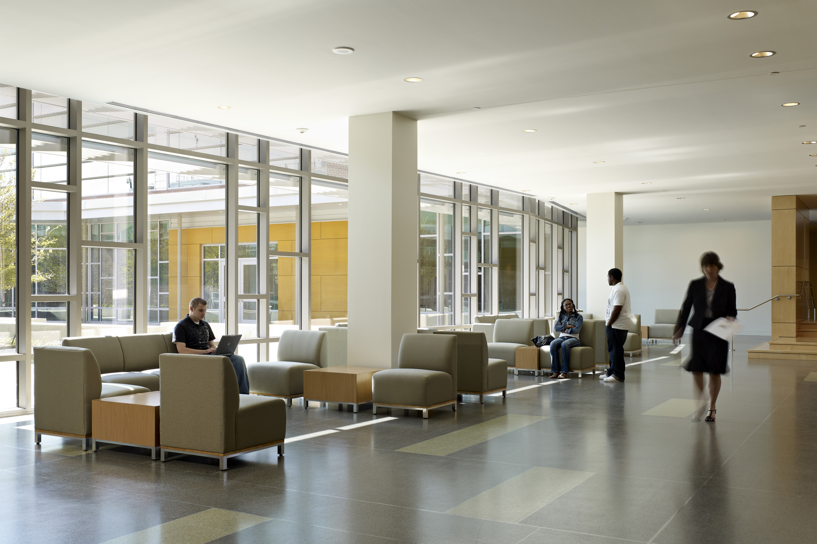 A spacious, modern indoor lobby with large windows, multiple seating areas, and a few people sitting and walking, reminiscent of the welcoming design found in Founders Hall at the University of North Texas.