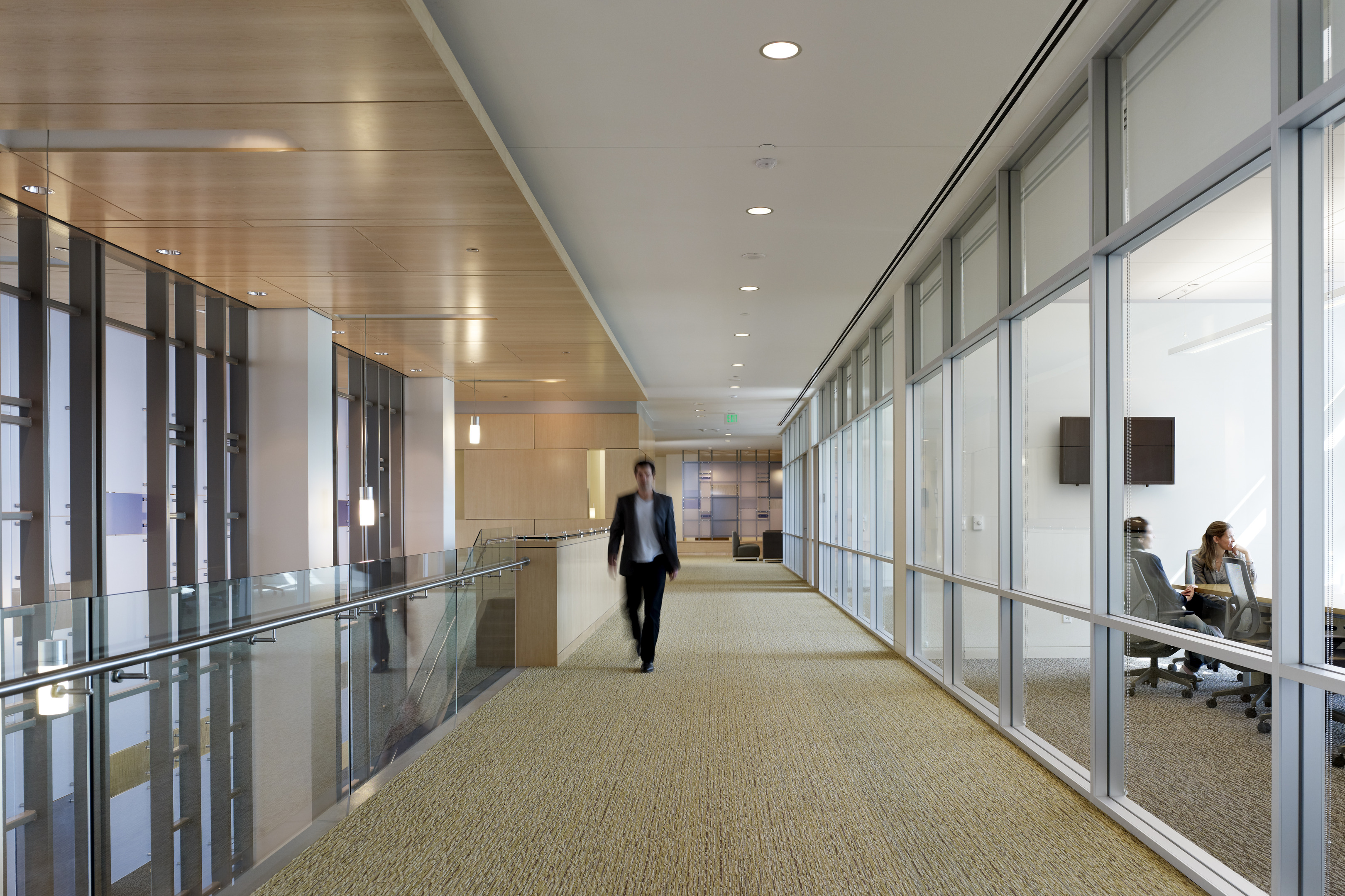 A person walks down a carpeted hallway in Founders Hall with glass walls on the right, through which two individuals can be seen seated and conversing in a meeting room at the University of North Texas.