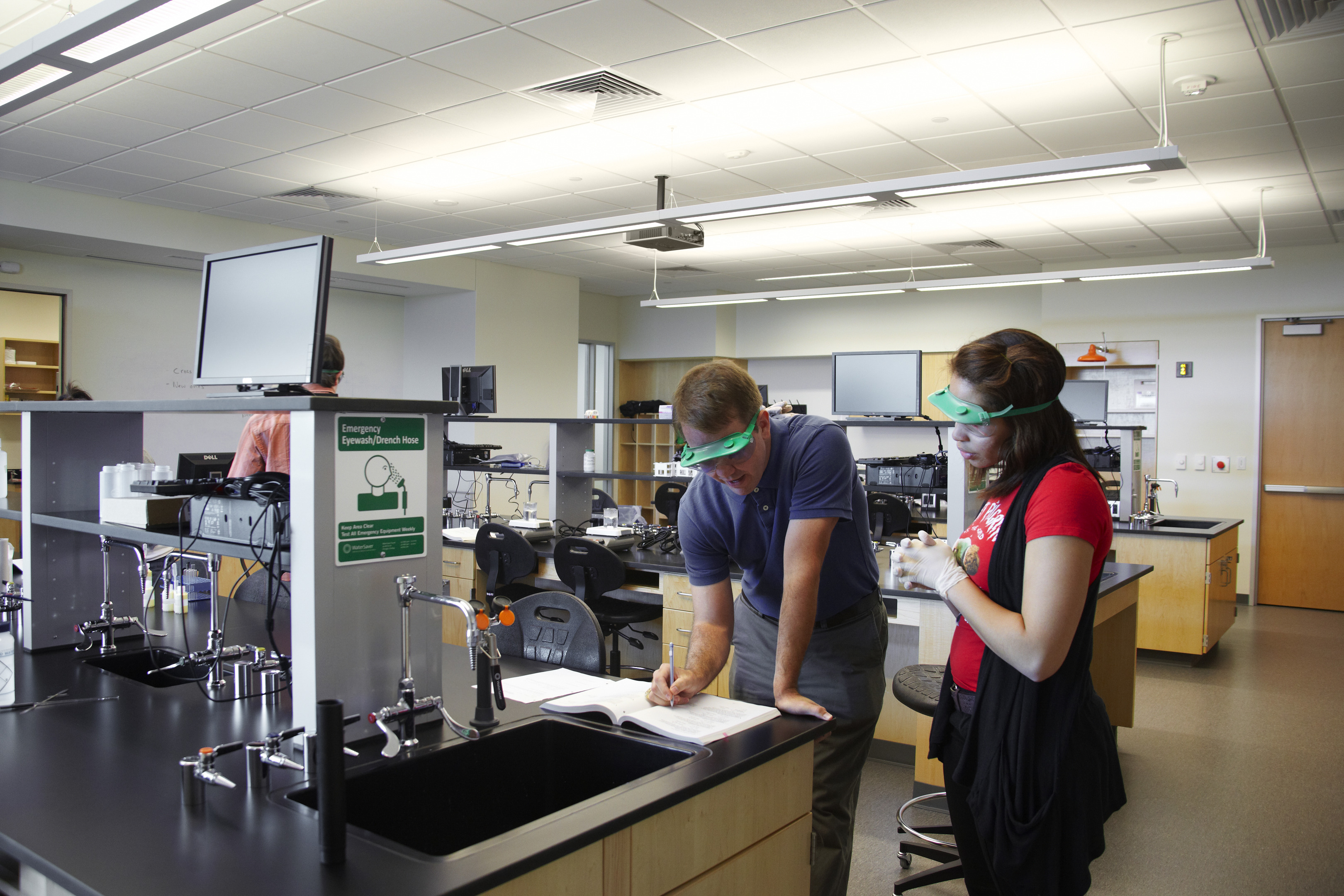 Two people wearing safety goggles are working together in a science laboratory at Founders Hall, University of North Texas. One is writing in a notebook while the other observes.