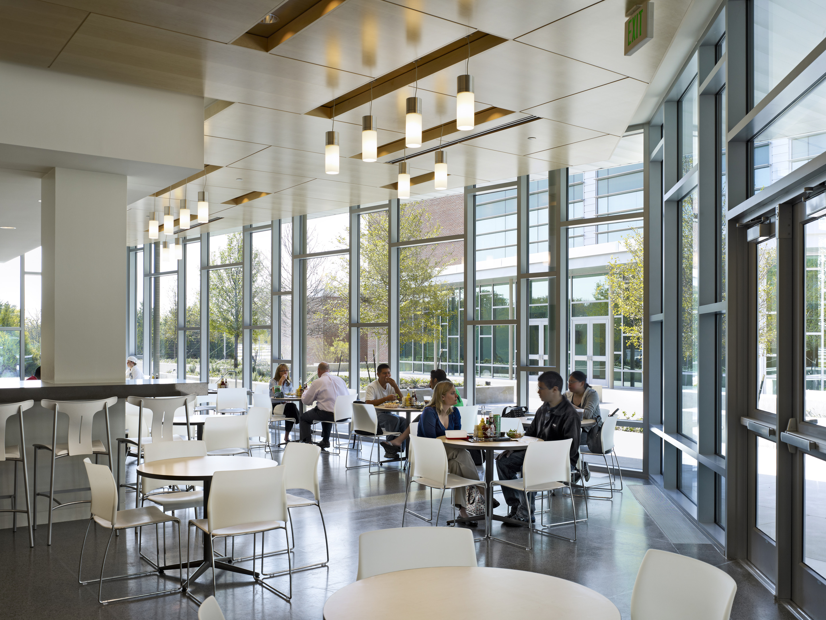 A modern cafeteria with large windows, round tables, and white chairs in Founders Hall at the University of North Texas. People are eating and conversing at various tables, creating a lively atmosphere reminiscent of Dallas' vibrant community spirit.