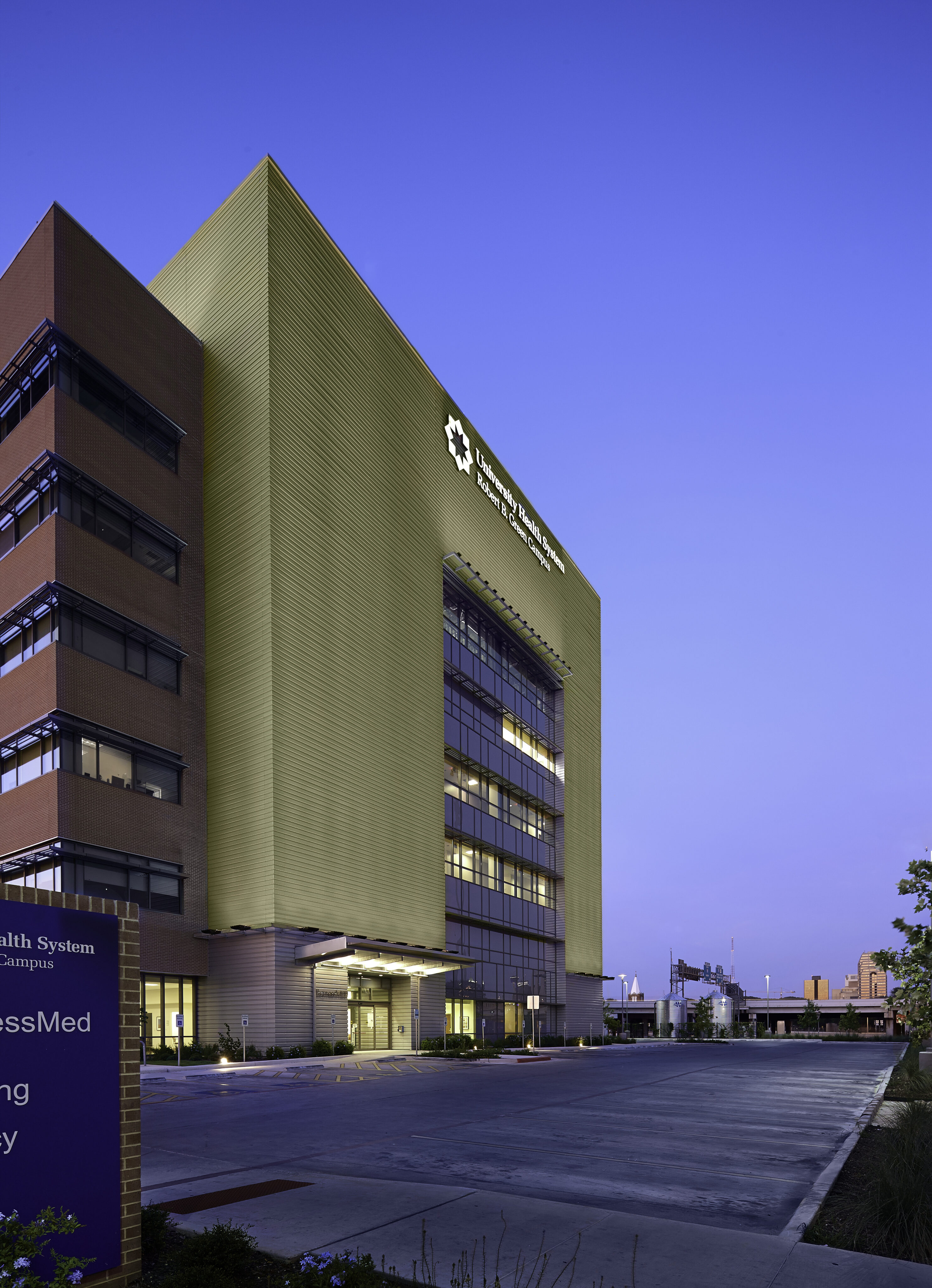 A tall, modern building made of glass and concrete stands against a twilight sky. The entrance and several floors are illuminated. Text on the building indicates it's the Robert B. Green Building related to the University Health System's medical campus.