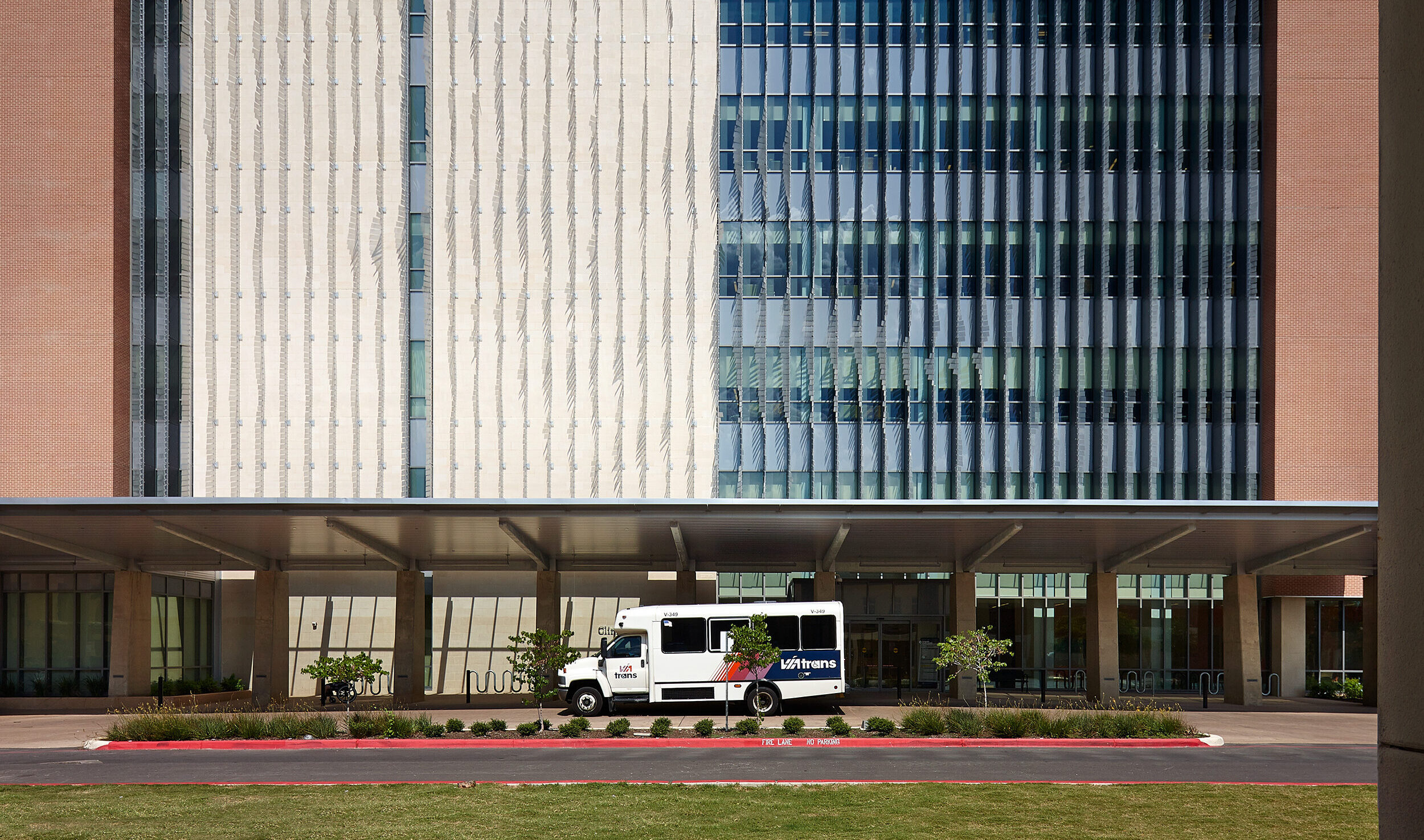 A white shuttle bus is parked in front of the Robert B. Green Building, a part of the University Health System, which features vertical window shades and a brick facade. A covered walkway extends from the building's entrance on this sunny day.