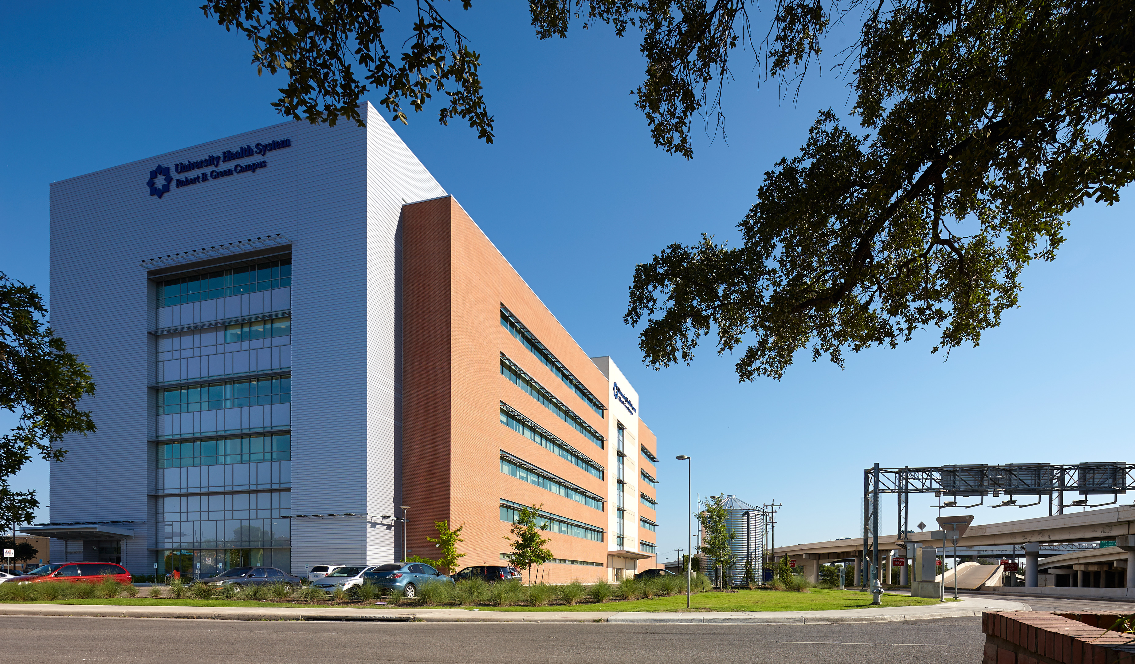 A multi-story hospital building, part of the University Health System, with a combination of glass, metal, and brick exterior under a clear blue sky, featuring surrounding trees and a nearby road with minimal traffic.