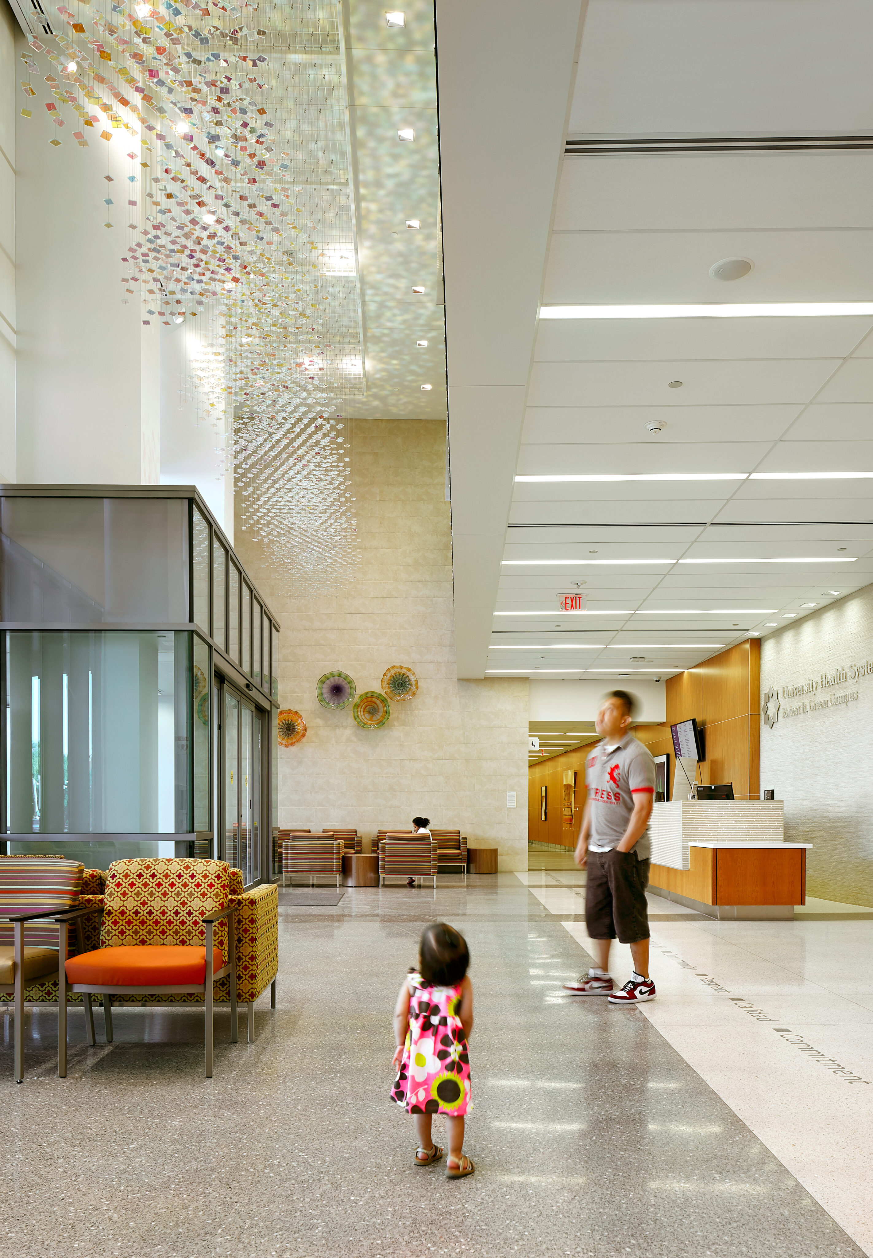 A man and a child stand in the modern lobby of the Robert B. Green Building, part of the University Health System, adorned with a high ceiling, colorful decorations, a seating area, and a reception desk in the background.