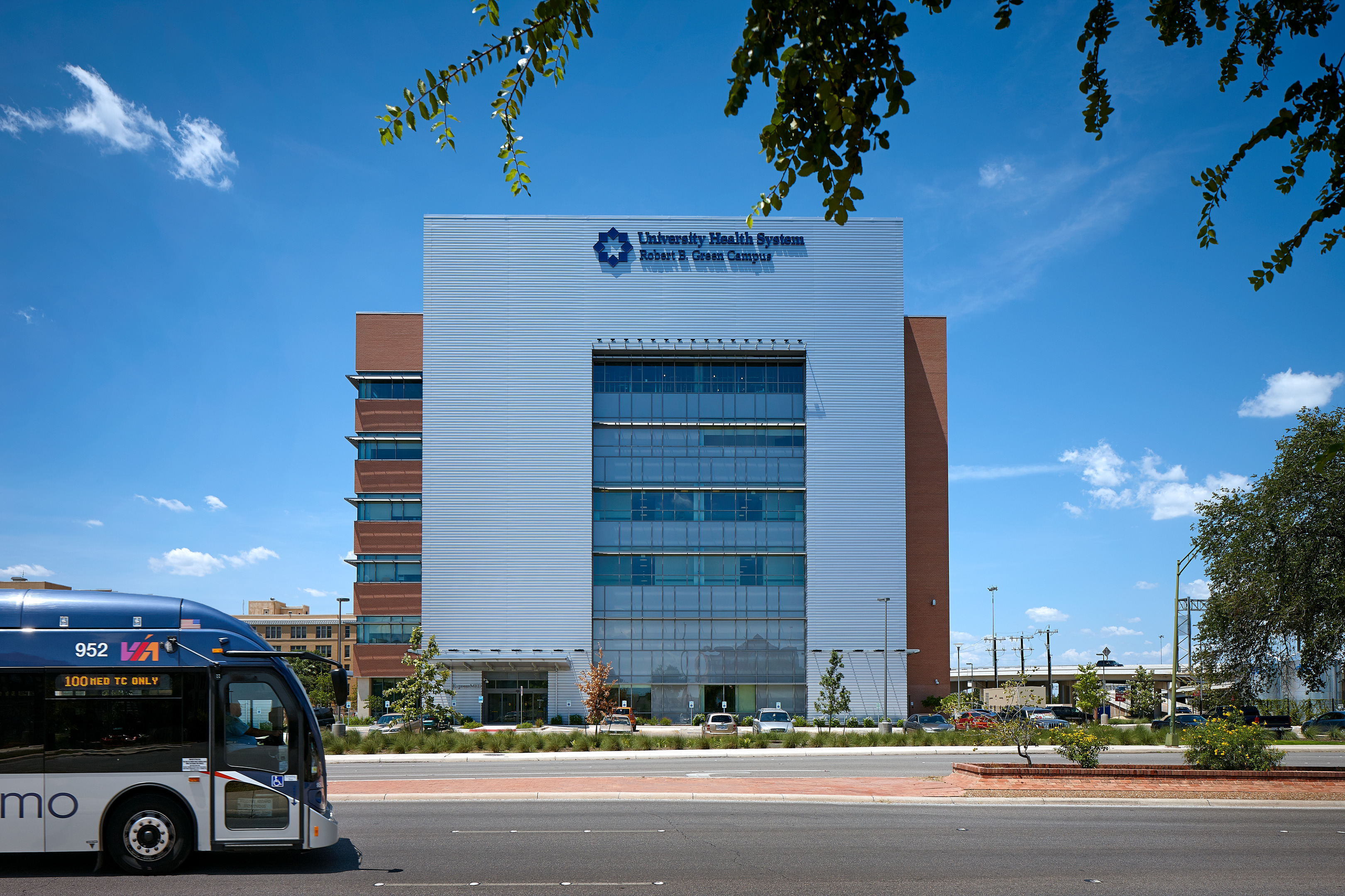 A six-story medical building with the sign "University Health System Robert B. Green Campus" in blue letters, seen from the street with a bus passing in the foreground. This landmark, known as the Robert B. Green Building, stands as a beacon of healthcare excellence.