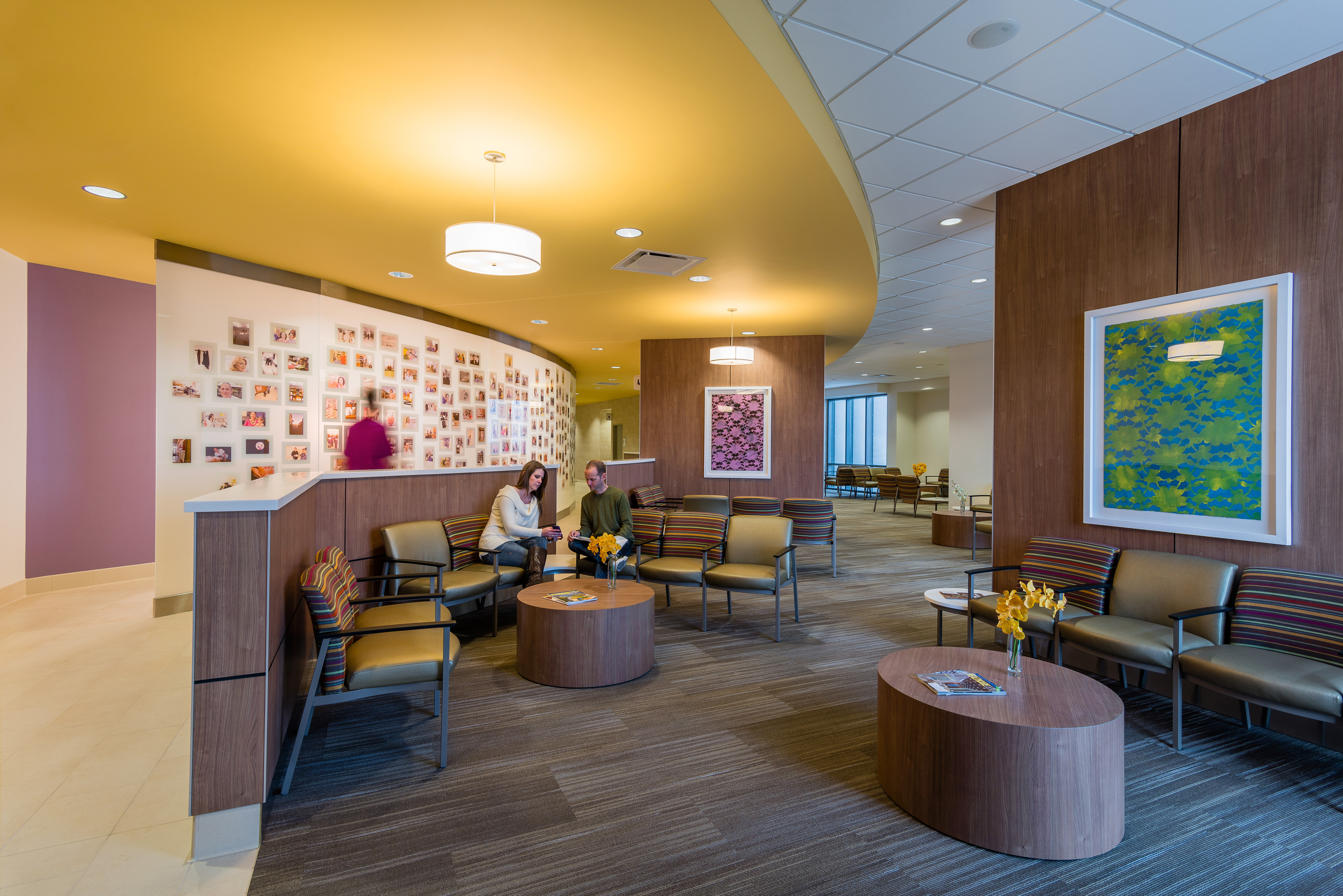 A modern waiting room in the Robert B. Green Building features a yellow ceiling, brown furniture, and wall art. Two people are seated and talking under framed photos displayed on a wall in the background, encapsulating the welcoming environment of University Health System.