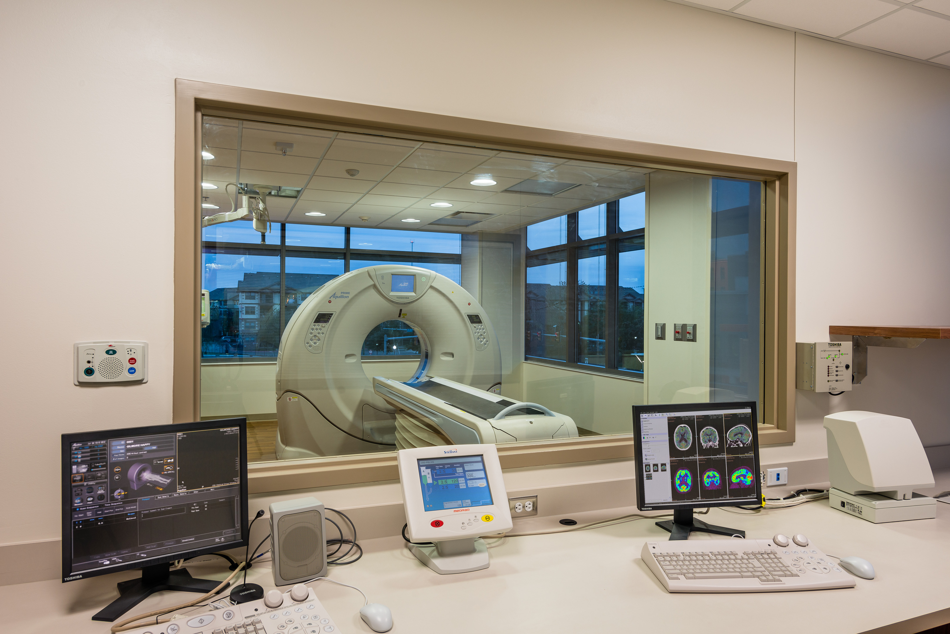 A medical imaging control room with monitors and control panels visible in the foreground, facing a CT scanner on the other side of a large window in the Robert B. Green Building at University Health System.