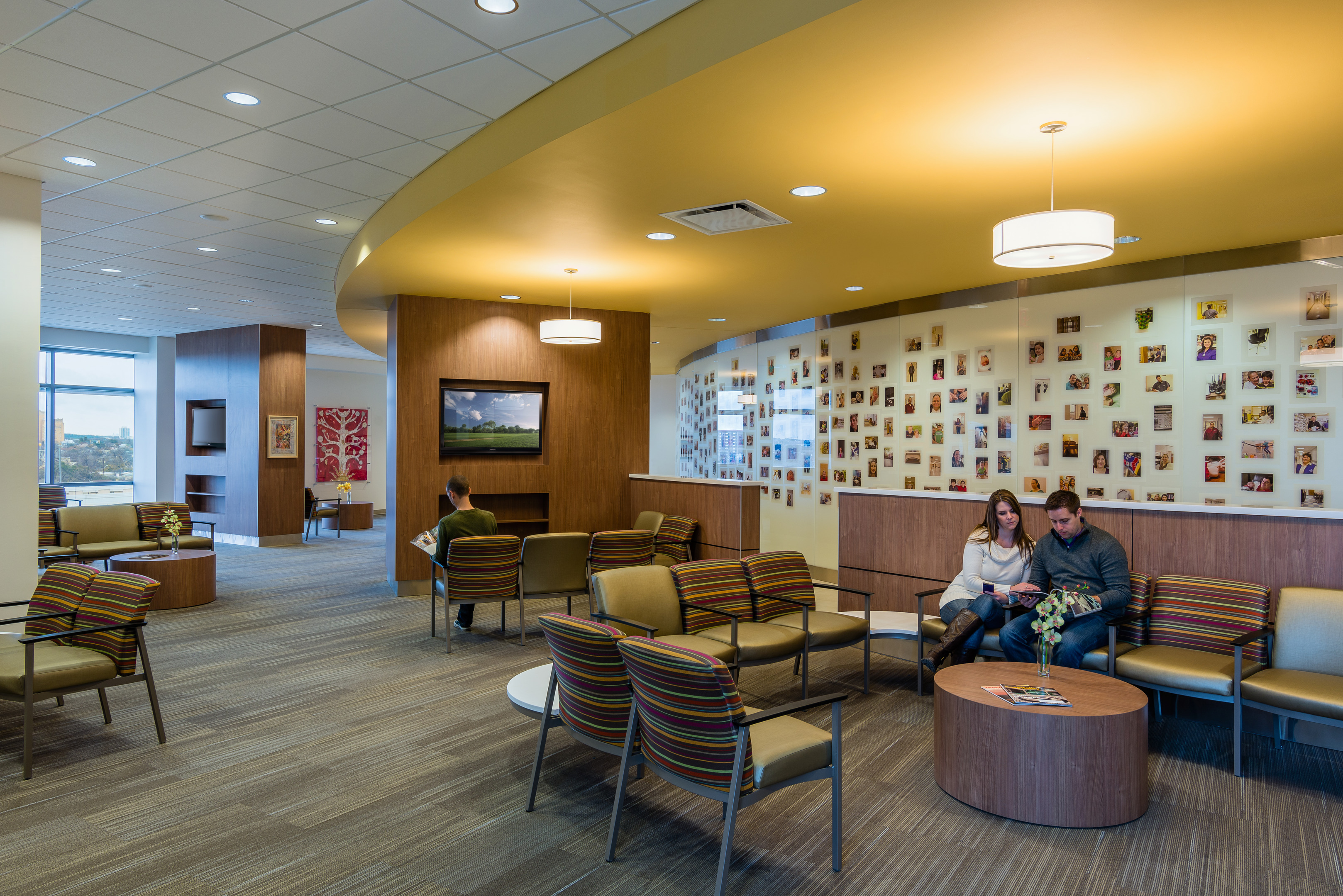 A waiting area in the Robert B. Green Building, part of the University Health System, features several chairs, a TV on the wall, and a yellow ceiling. People are seated; a man alone and a couple looking at a magazine. The wall is adorned with numerous small photos.