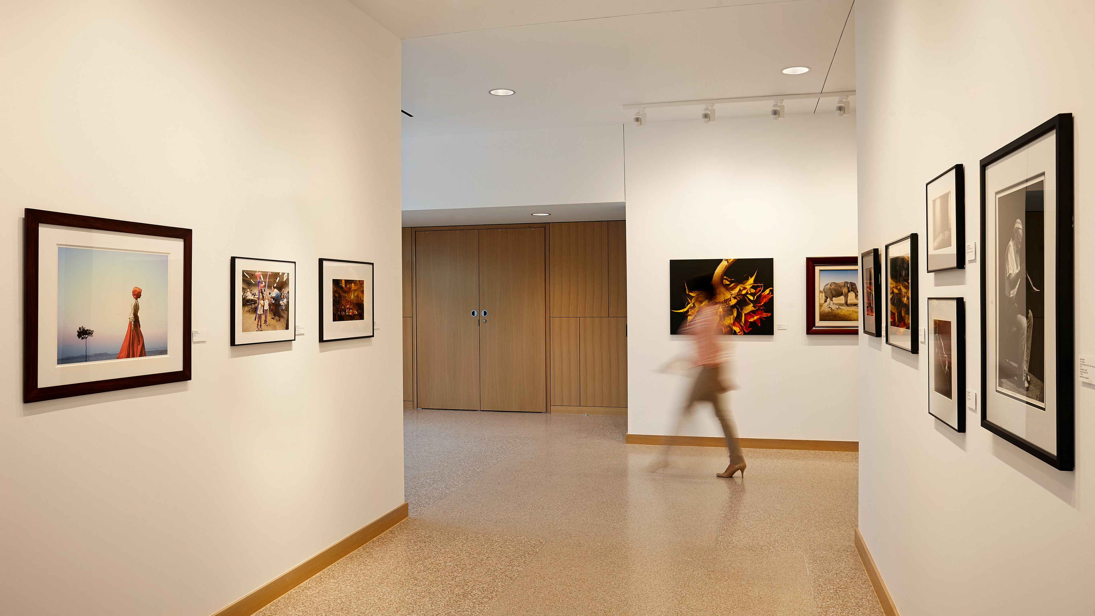 A person walks past framed photographs and artwork displayed on the walls of a modern, well-lit gallery at Rice University's Anderson-Clarke Center.