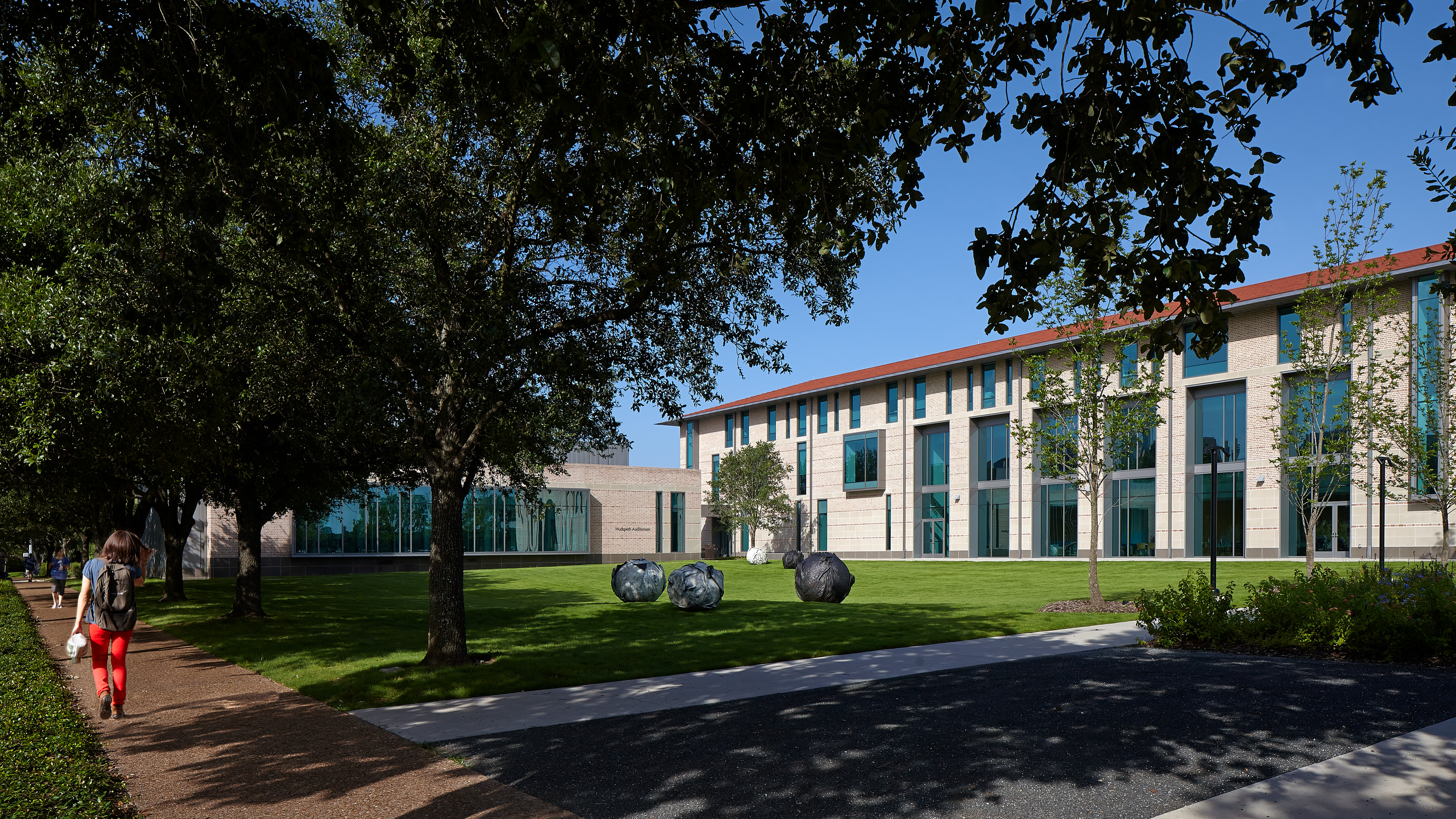 Green lawn with several large rocks in front of the Anderson-Clarke Center at Rice University, a modern building with lots of windows. A person with a backpack walks along a path shaded by trees on the left. Clear blue sky above.