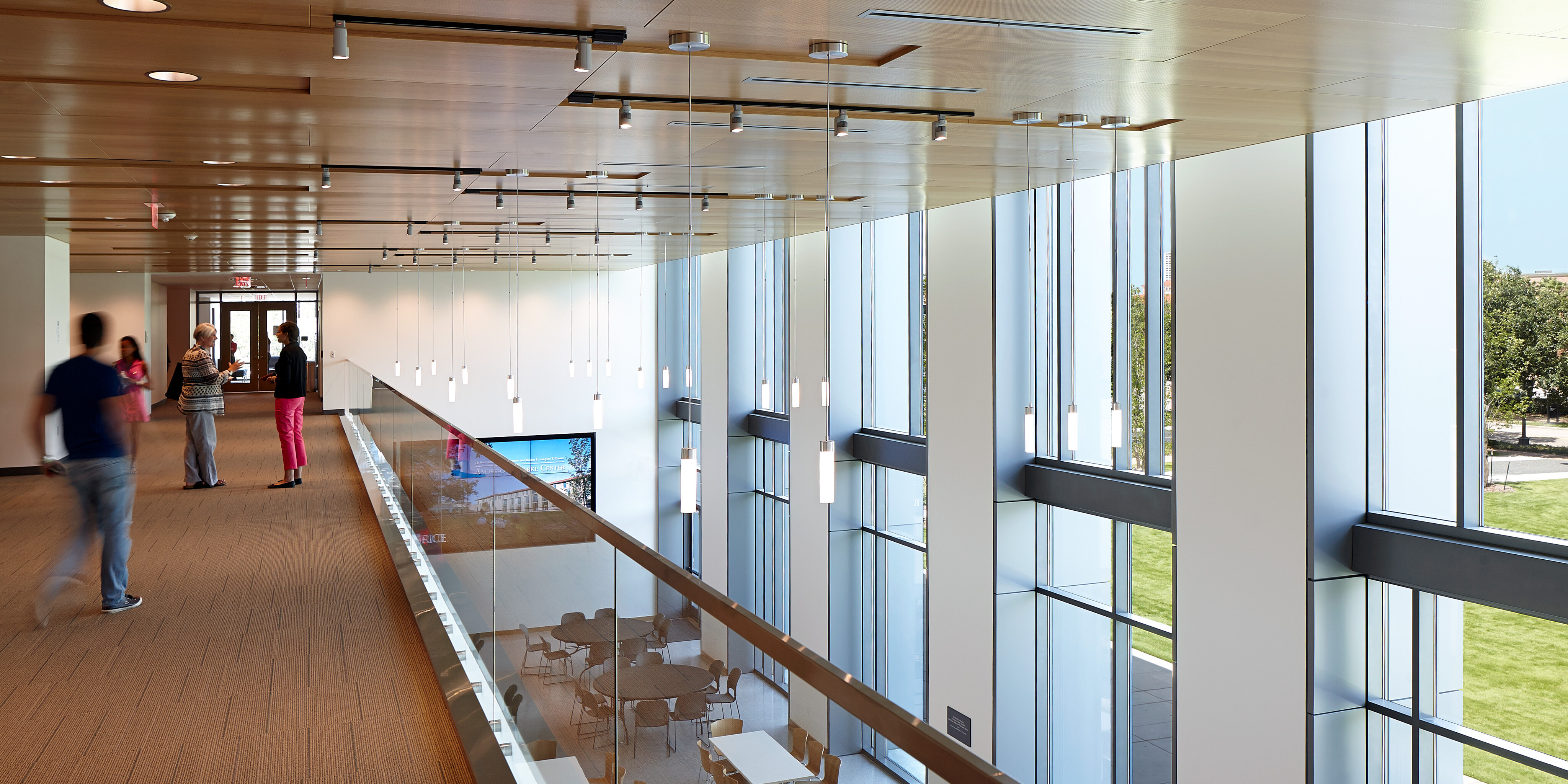 A spacious, modern hallway at the Anderson-Clarke Center, with large windows overlooking a green area at Rice University. A few people are walking and talking, and several small tables and chairs are visible on the lower level.