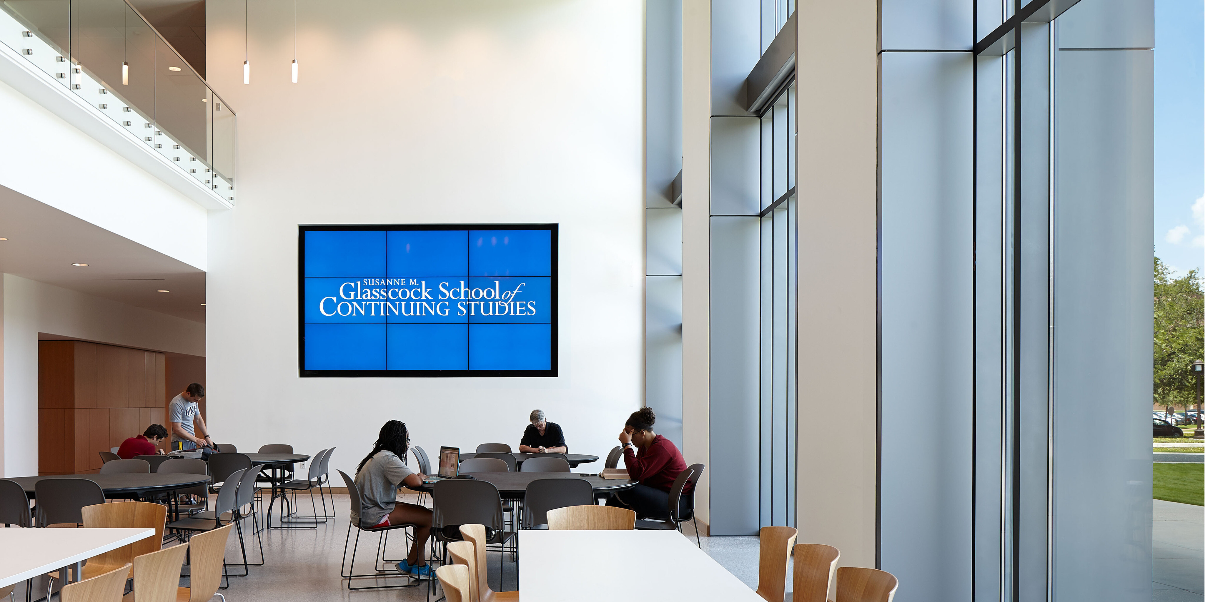 People sitting and studying at tables in a bright, modern room with large windows at Rice University's Anderson-Clarke Center. A large screen on the wall displays "Glasscock School of Continuing Studies.