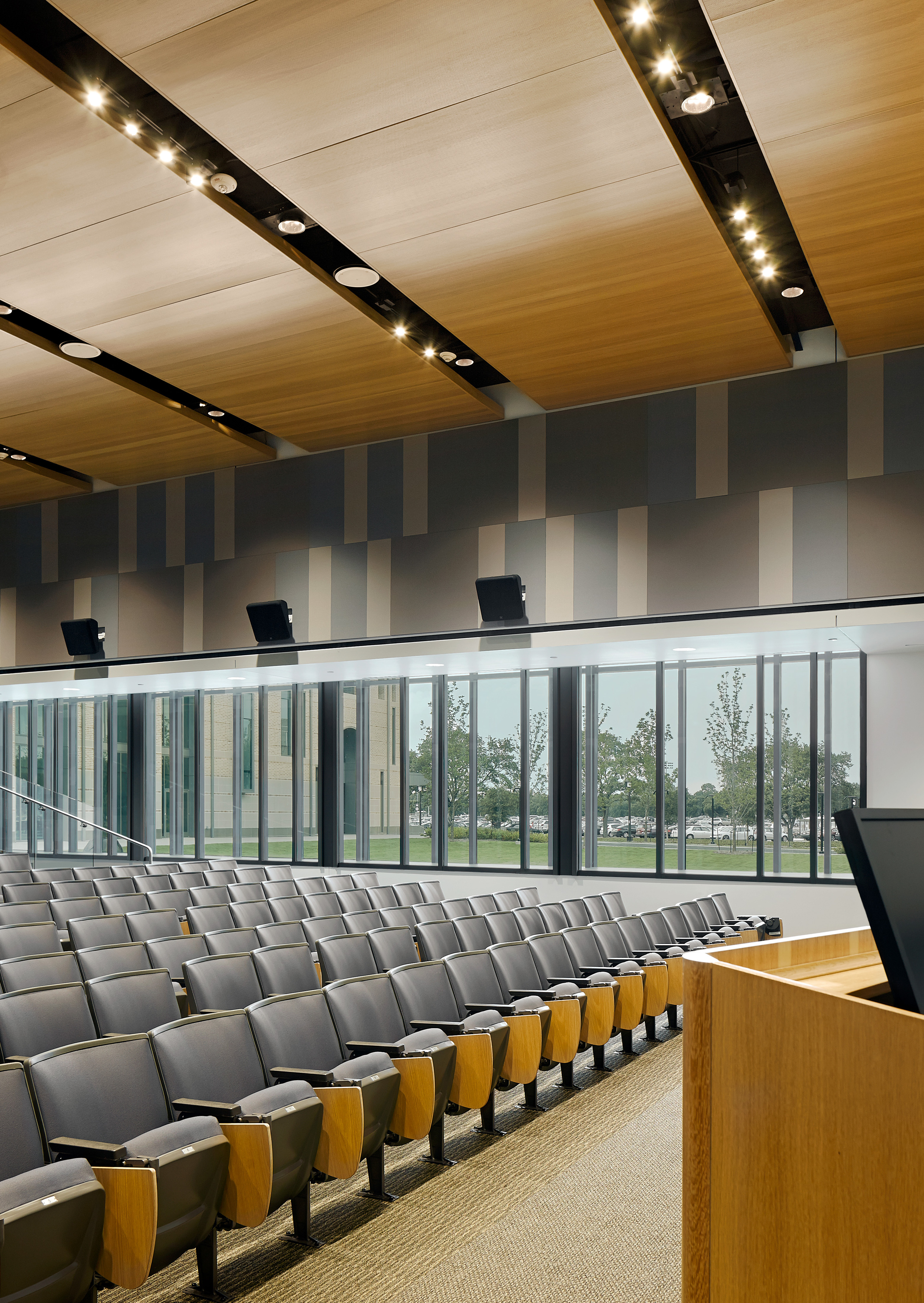 An empty lecture hall with rows of gray seats, a wooden podium, and large windows offering a view of Rice University’s Anderson-Clarke Center surrounded by trees and buildings.