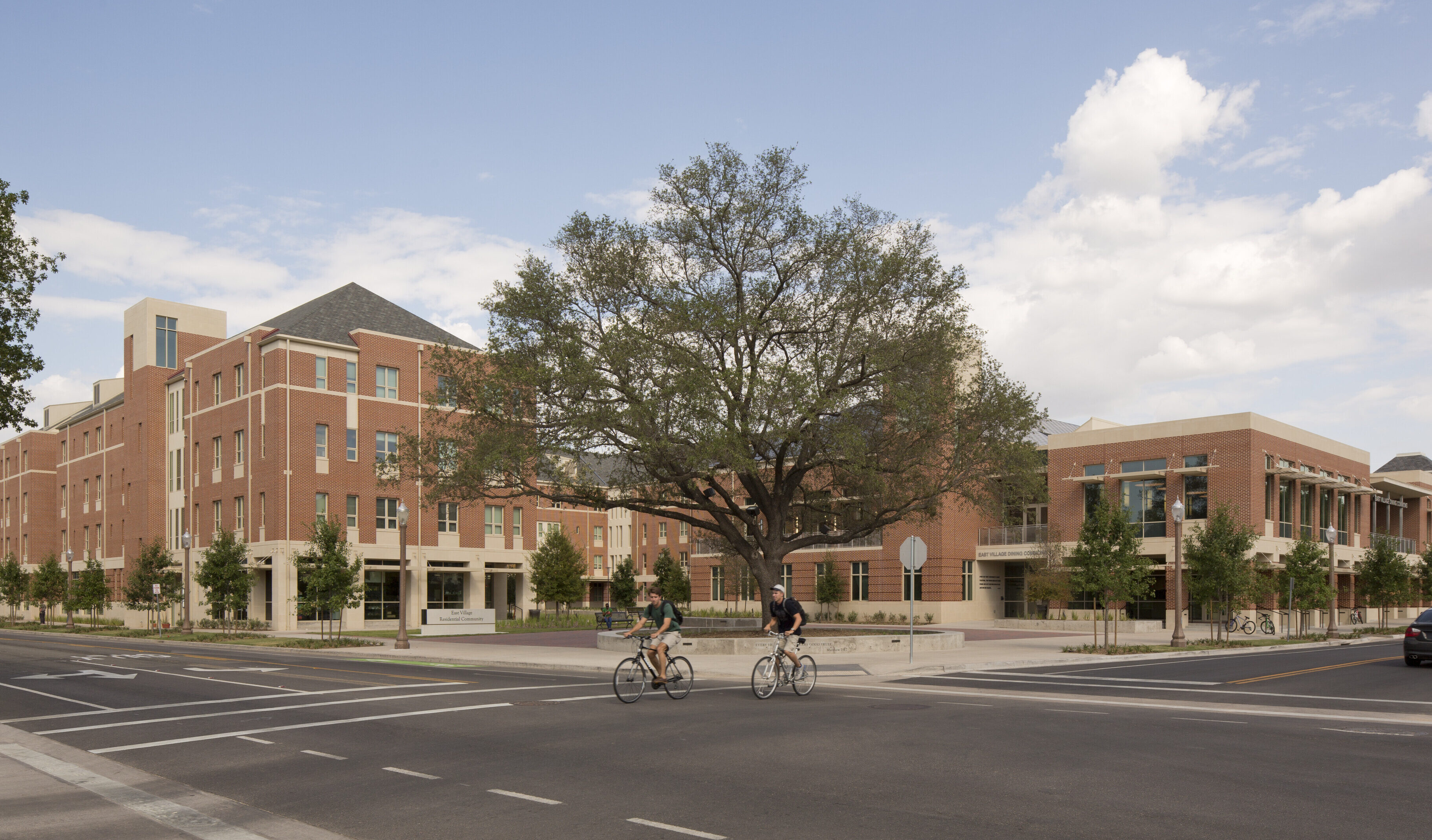 A large, tree-lined academic building with red brick walls stands at a street intersection in Baylor's East Village, with two cyclists riding in front under a blue sky with scattered clouds.