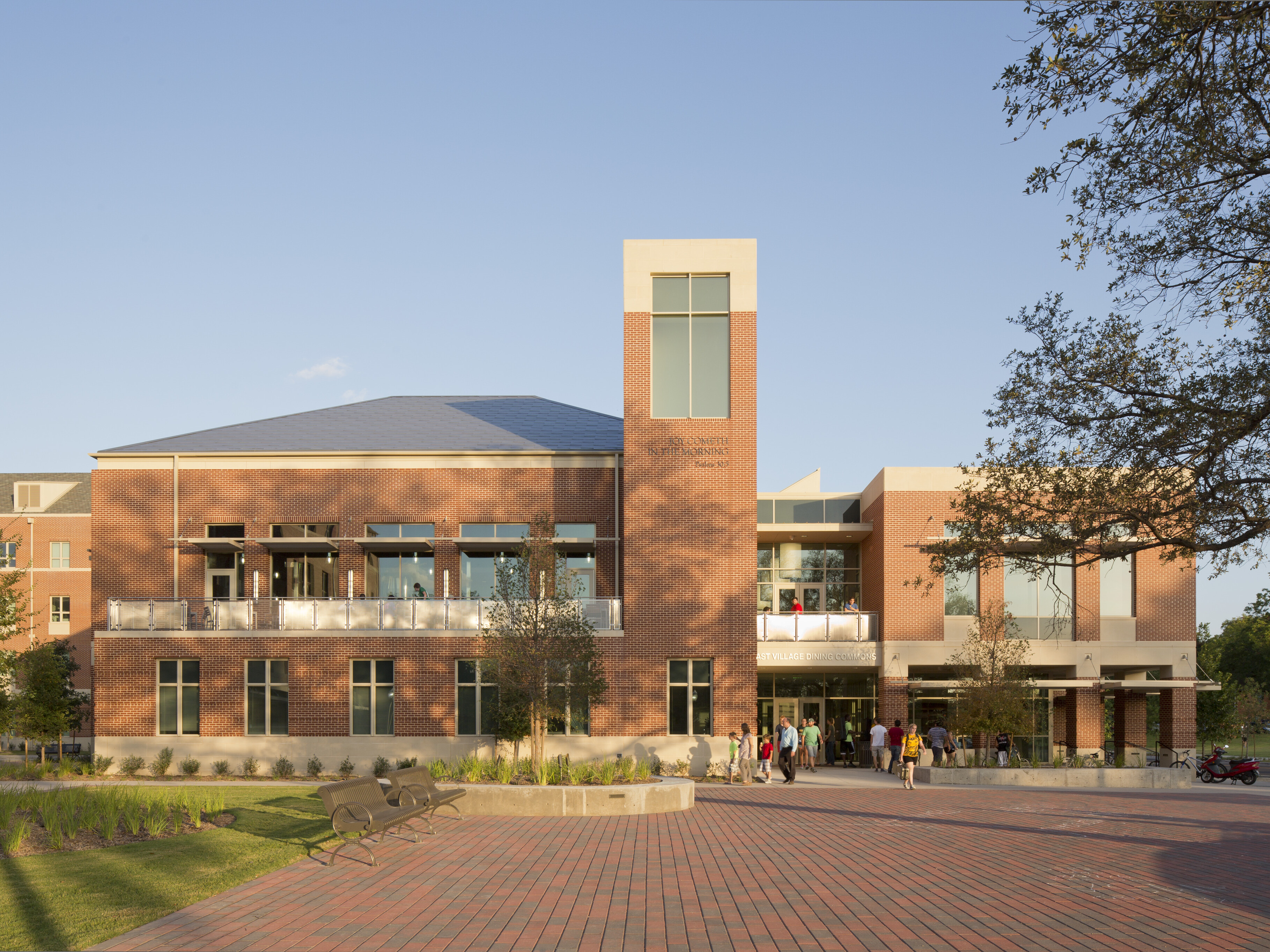 A modern brick building with large windows and a tall section labeled "Smith Building," located in East Village, serves as a student residence for Baylor, surrounded by a paved plaza, benches, and trees under a clear sky.