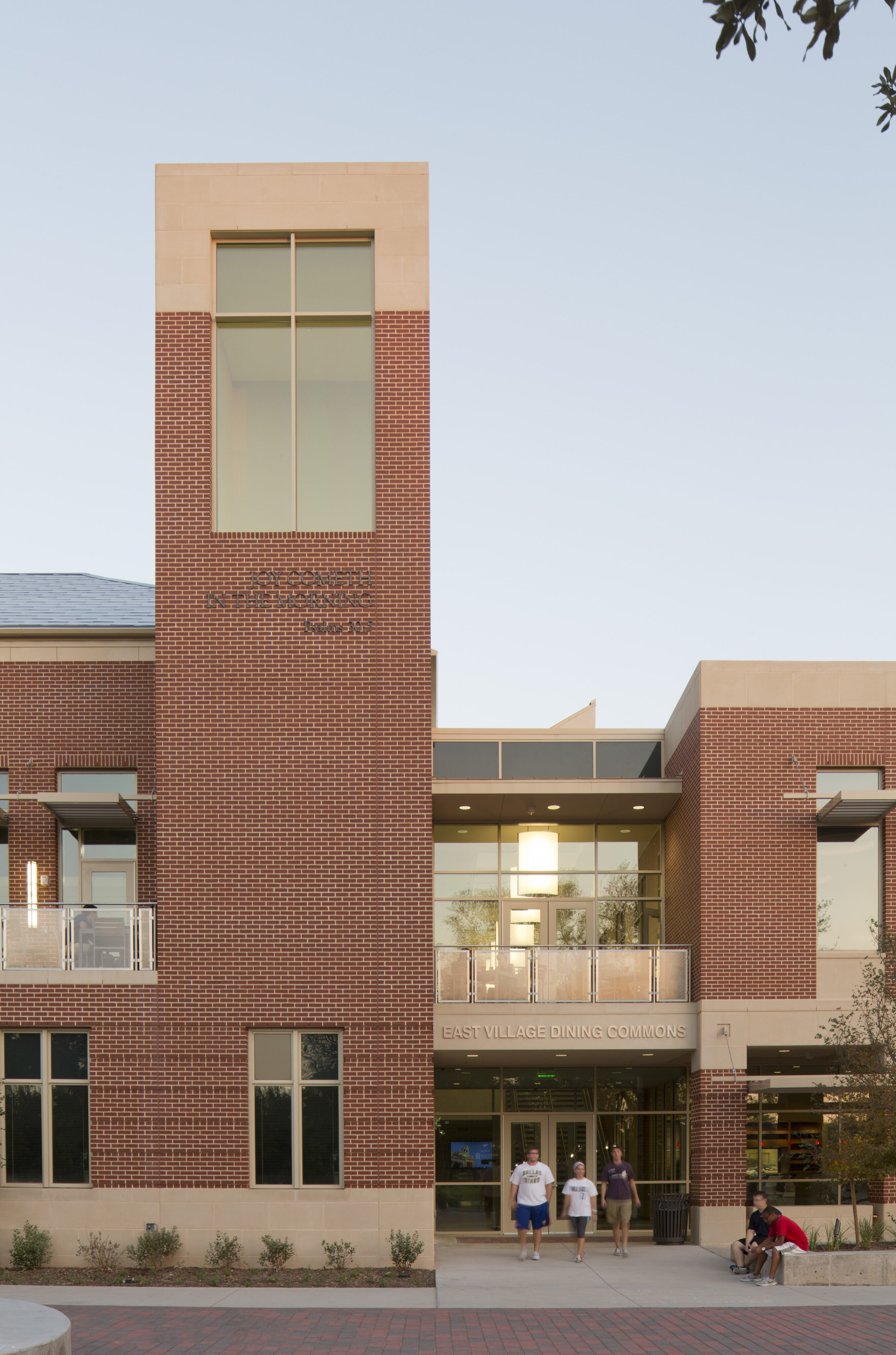 A brick and glass building with a tall central tower labeled "Solomon Residential College" and "East Village Dining Commons." Several people, likely Baylor students, are walking in front of the entrance.