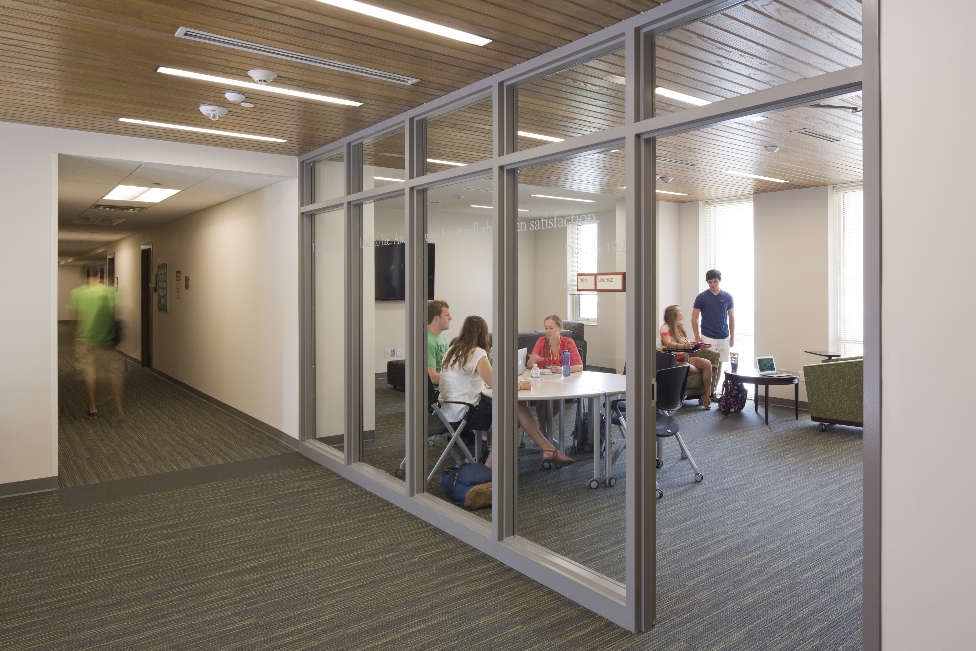 A group of people is having a meeting in a glass-enclosed room at the East Village Student Residence, with a person walking down the adjacent hallway.