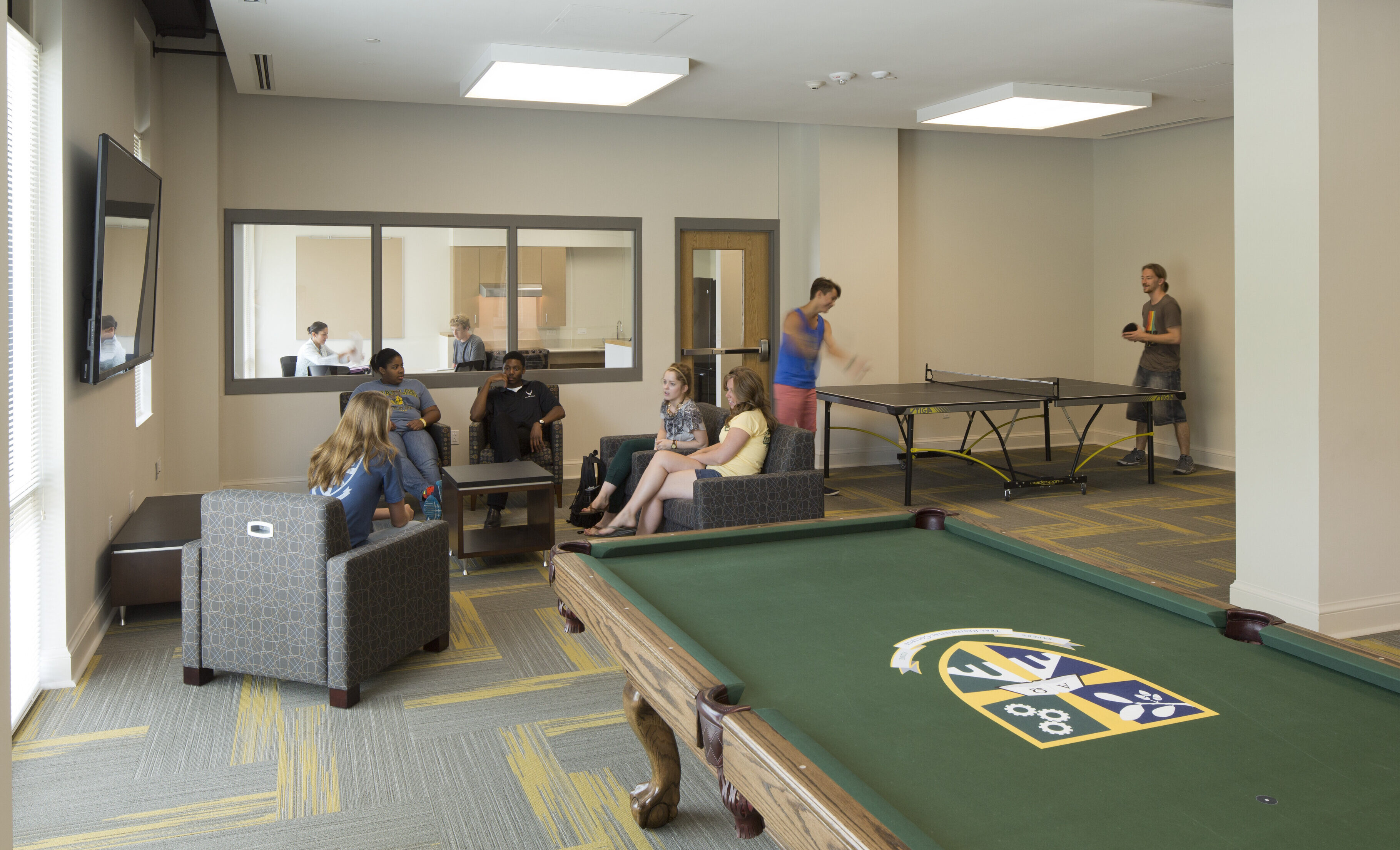 In the East Village Baylor student residence, people are gathered in a communal lounge area with a pool table, TV, sofas, and a ping pong table. Some are seated talking while others are playing. A group is in a room visible through a window.