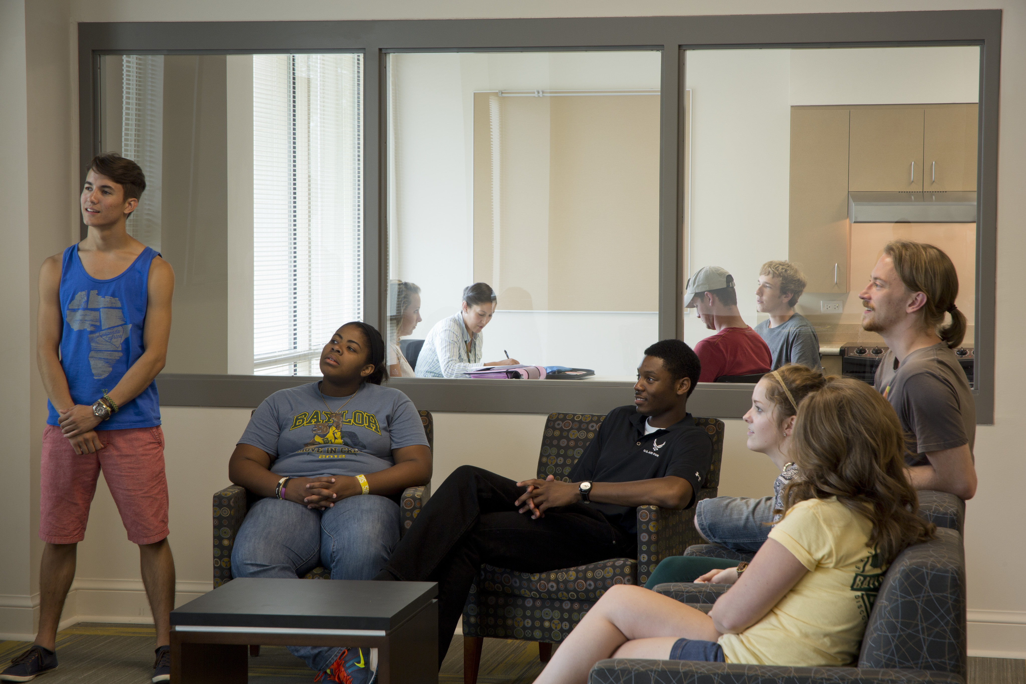 A group of people, possibly from Baylor, are sitting and standing in a common area having a conversation. Six individuals are visible, with some seated on chairs and others standing, in front of a window looking into another room in the East Village Student Residence.