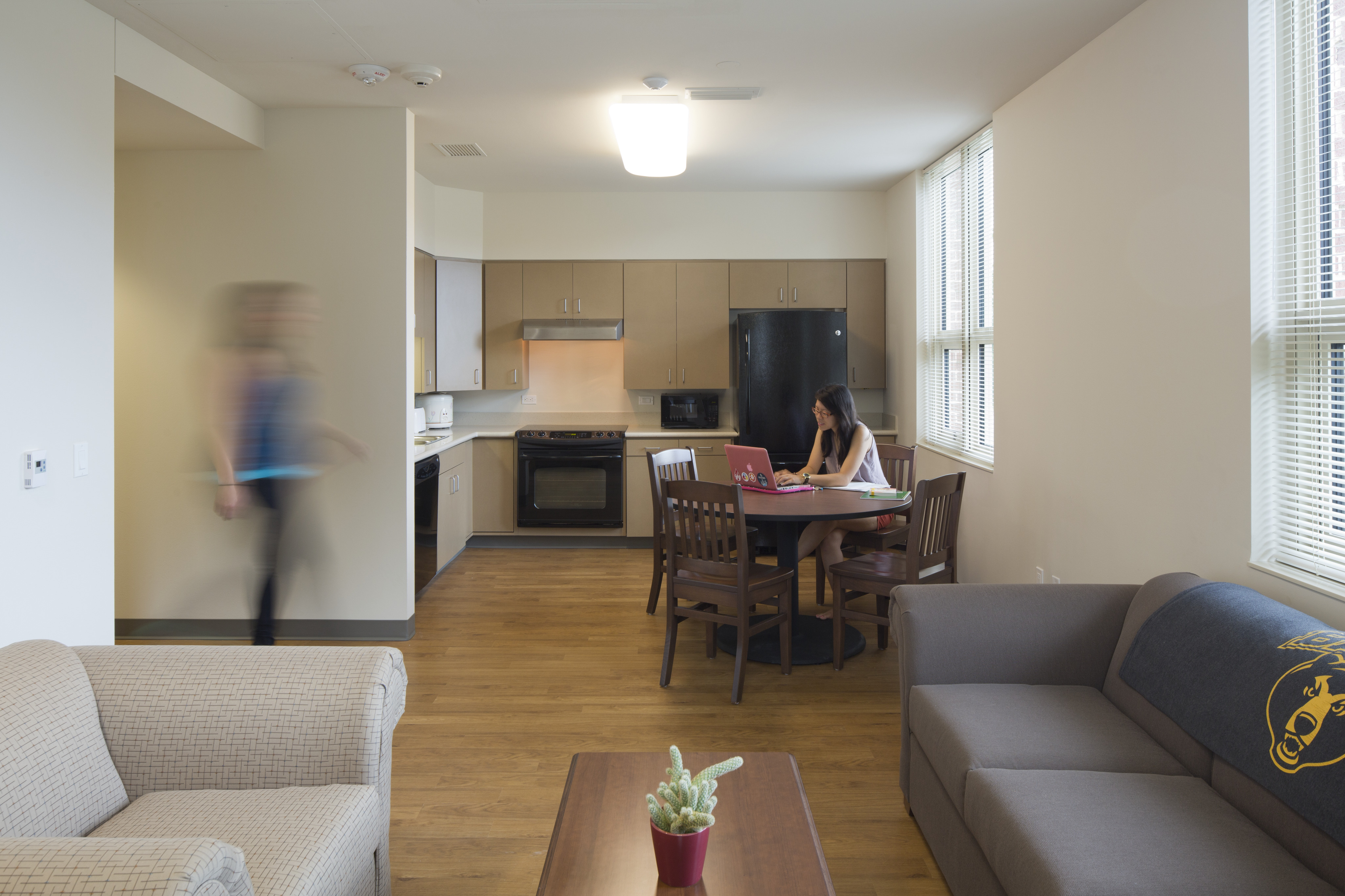 A person is seated at a table using a laptop in a modern kitchen and living area, typical of a Baylor student residence in East Village. Another person is walking past in a blur. The room is well-lit with wooden floors, beige walls, and contemporary furniture.