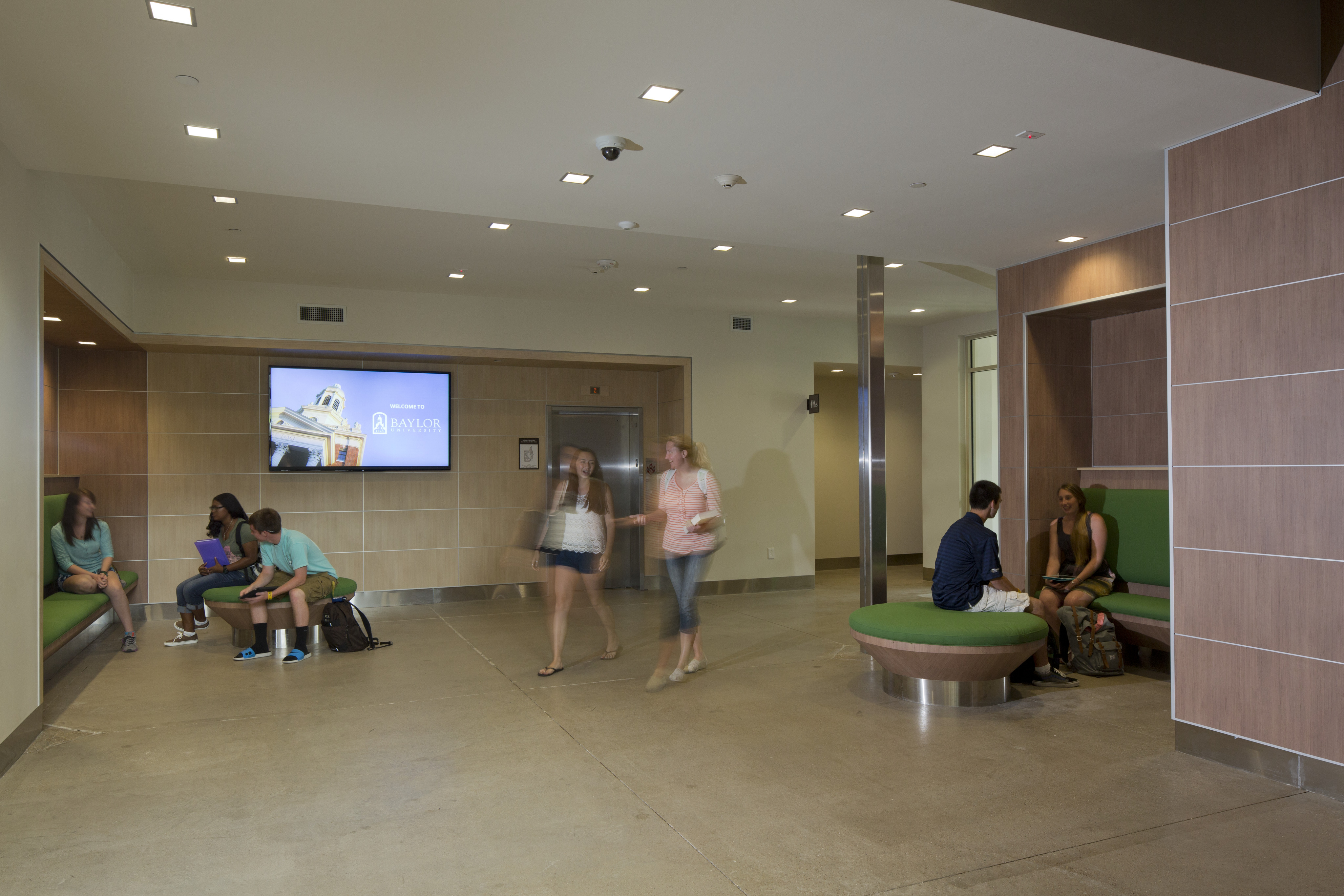 A lobby area in the Baylor Student Residence with people sitting on benches and a round seat, a digital screen displaying "Randall Pavilion," and two individuals walking past.