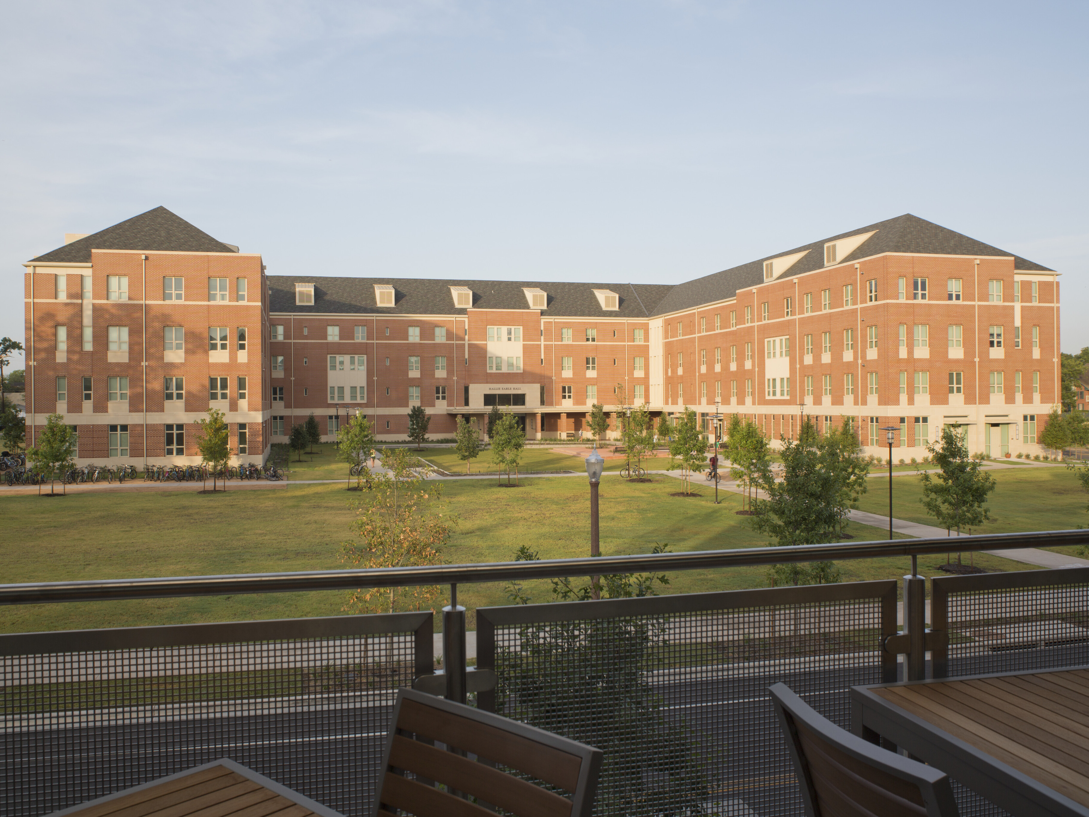 A three-story brick academic building with multiple windows and peaked roofs, surrounded by a grassy lawn and trees, is viewed from a terrace with wooden tables and chairs—reminiscent of the serene setting of Baylor's East Village Student Residence.