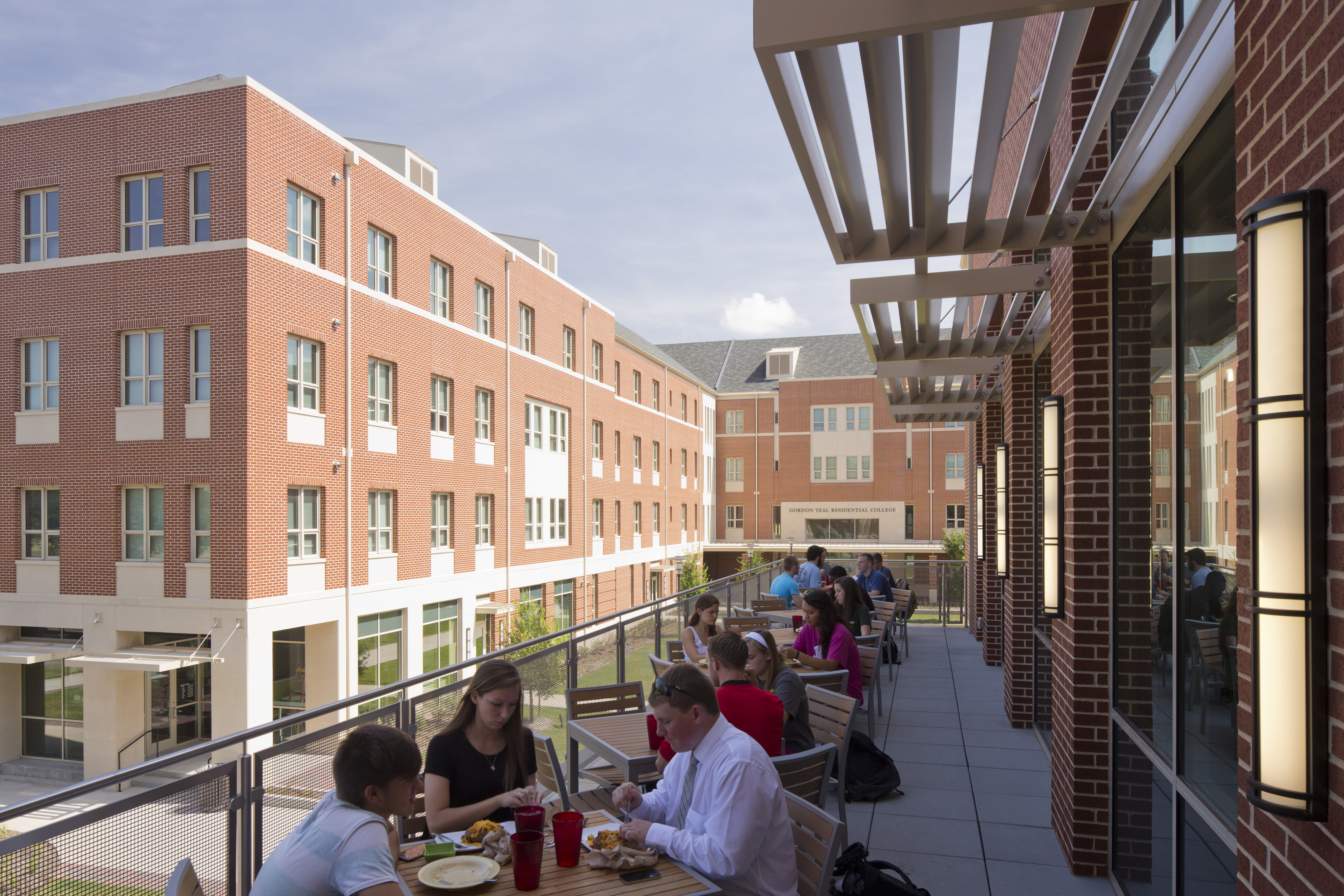 People seated at tables on an outdoor patio in East Village, eating and conversing, with multi-story brick buildings and the Baylor Student Residence in the background under a clear sky.