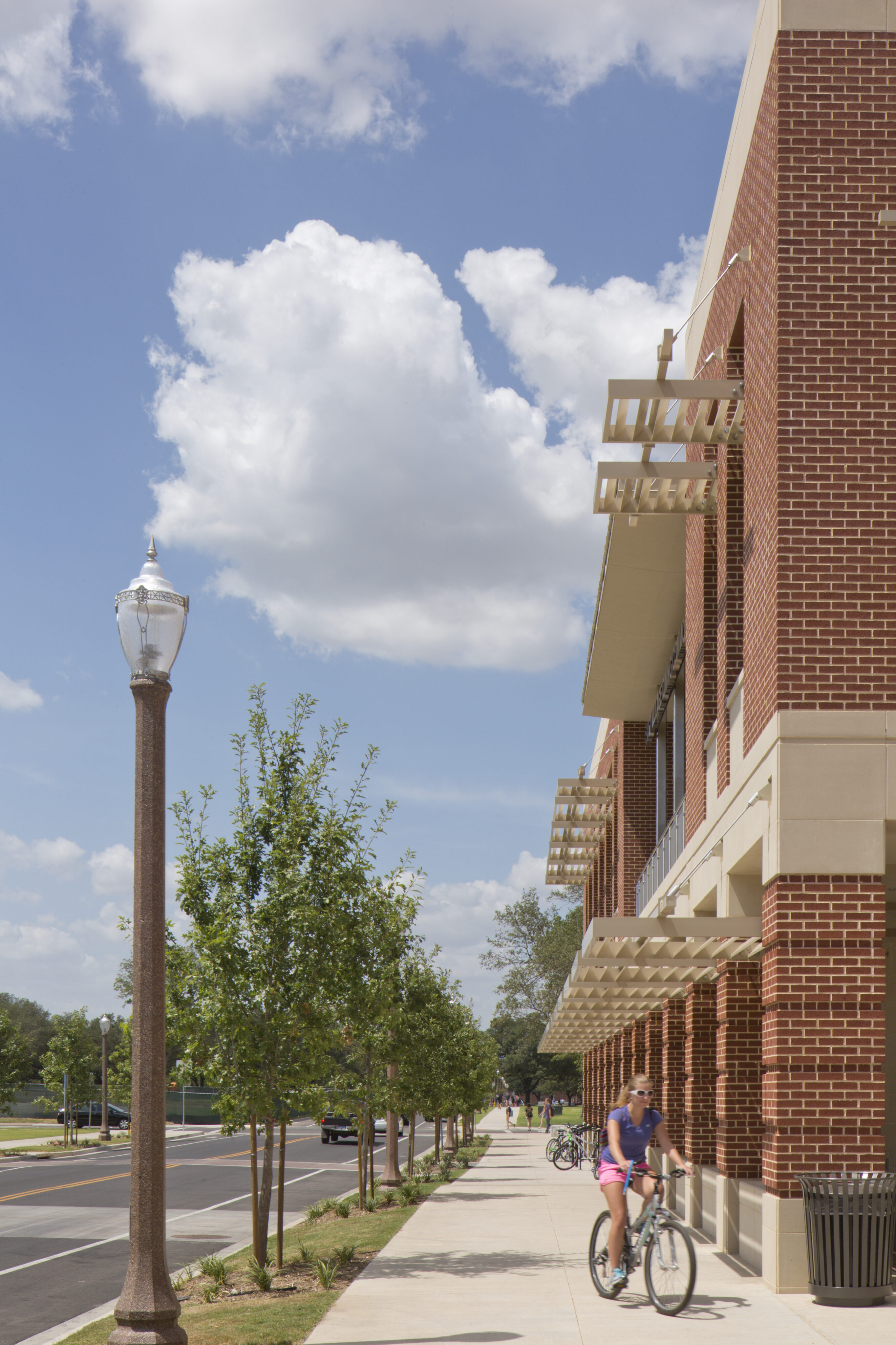 A person rides a bicycle on the sidewalk next to a modern brick building with awnings in East Village under a partly cloudy sky. Street lamps and young trees line the path, leading to student residences at Baylor.