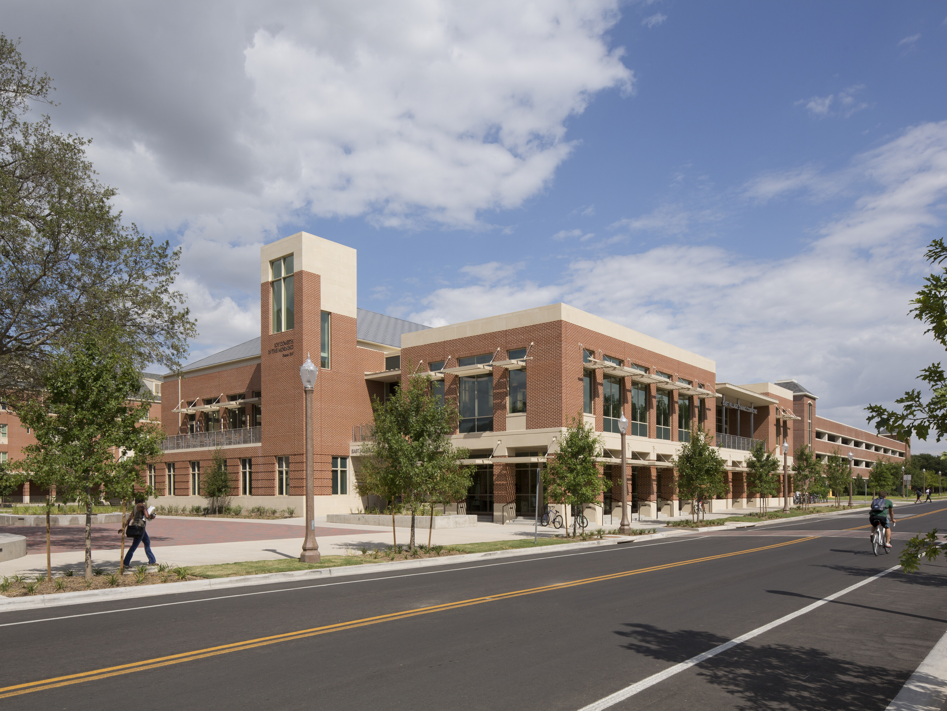 Two-story brick and stone building with large windows, surrounded by trees in East Village. People are walking and cycling on the street under a partly cloudy sky, giving the area a vibrant community feel. This charming student residence offers a perfect setting for Baylor students.