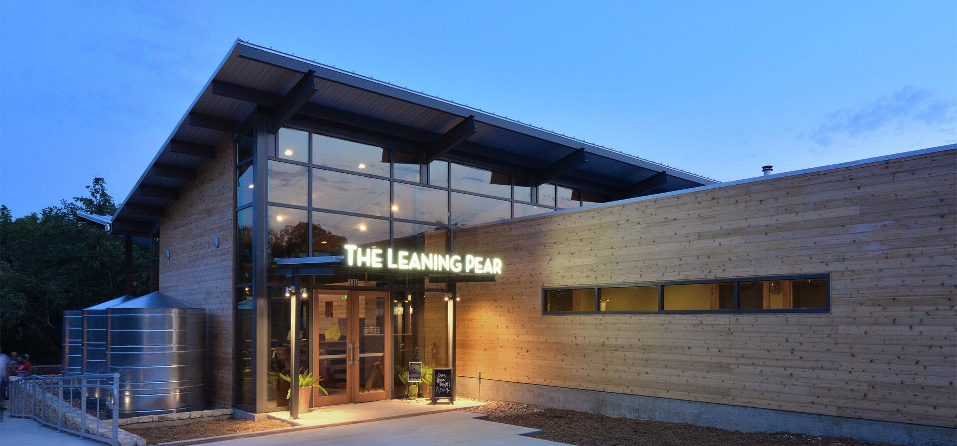 Exterior view of The Leaning Pear restaurant at dusk showcases a modern wooden design, large windows, and the name illuminated above the entrance. The auto draft ambiance of twilight enhances its inviting allure.