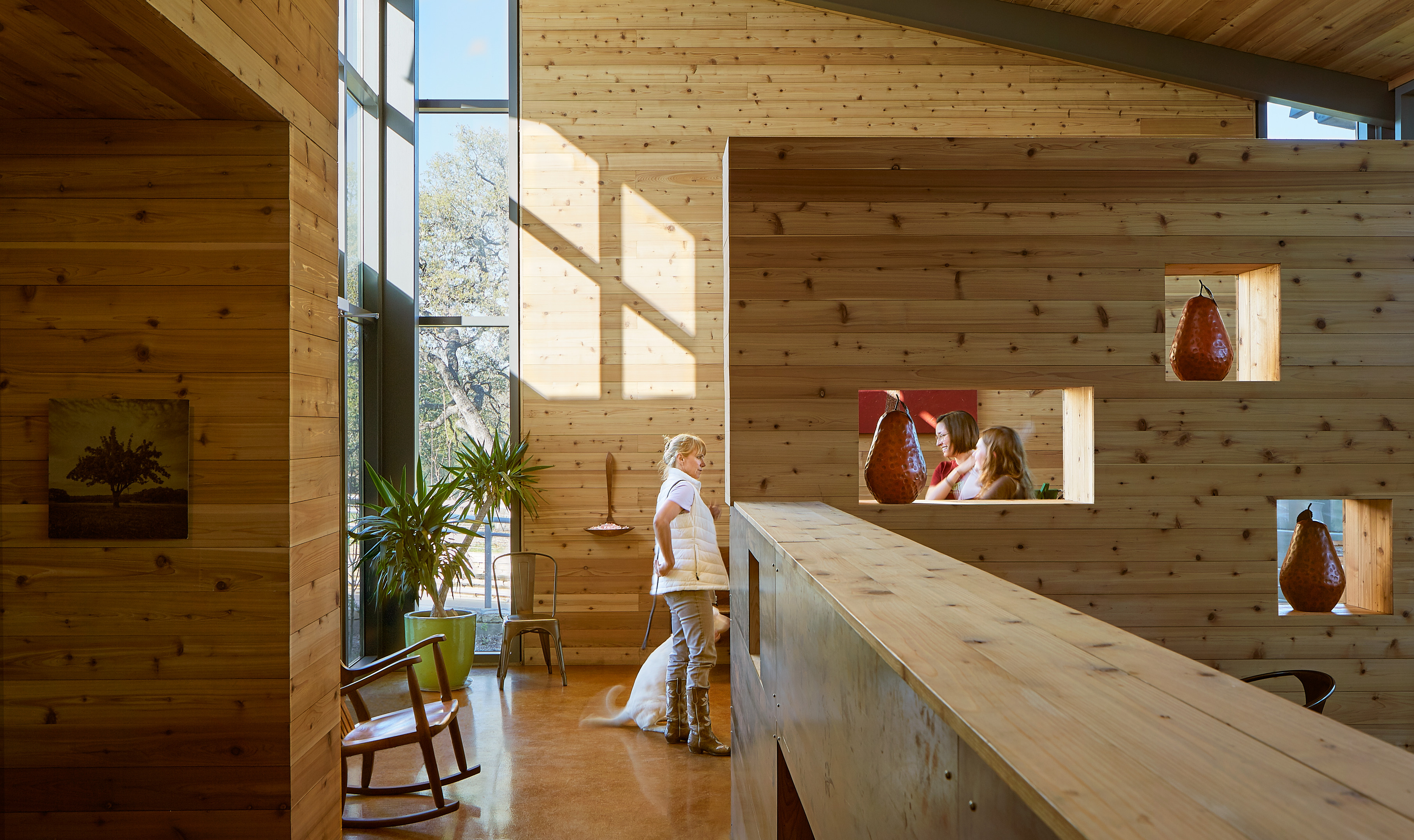 A person and a dog are in a wooden interior with large windows. Through a wall opening, two women can be seen talking. The space features wooden furniture, vases displayed in built-in niches, and has an effortless charm reminiscent of an Auto Draft design.