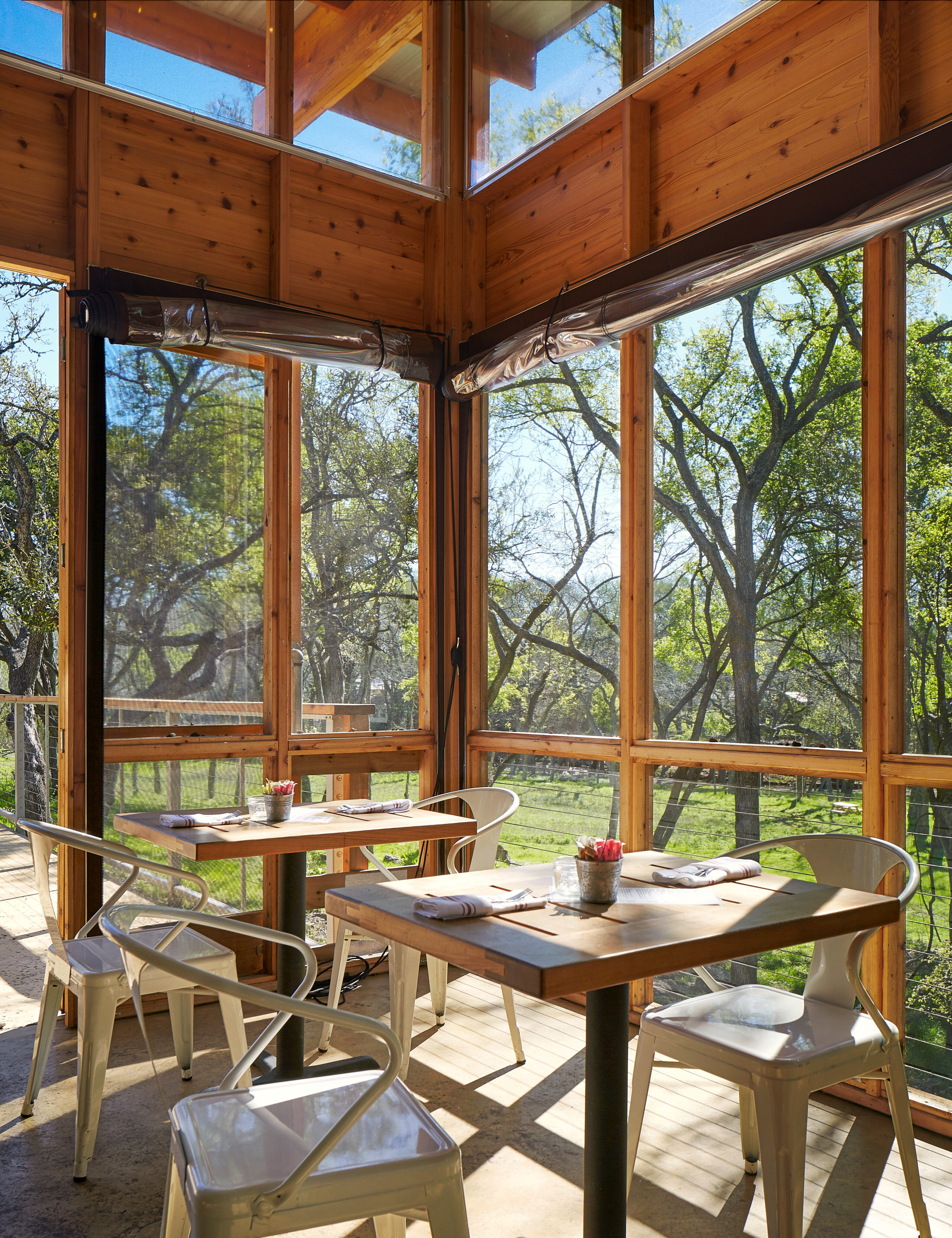 A sunlit room with large windows features two tables, each set with chairs and dining utensils, overlooking a green, tree-filled landscape. The serene ambiance feels like an Auto Draft designed by nature itself.